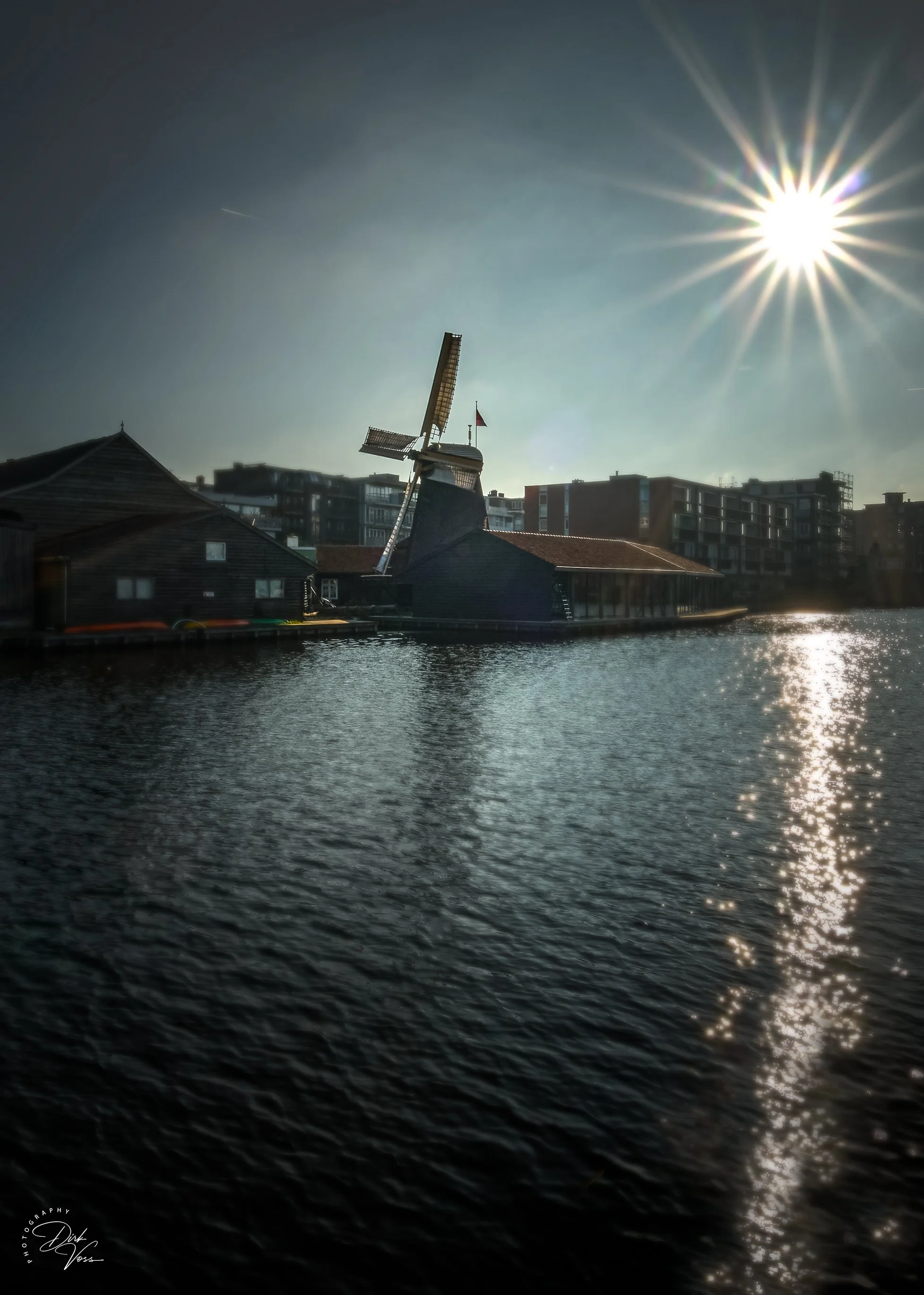 Ein Kanal mit Häusern am Ufer, eine Windmühle und moderne Gebäude im Hintergrund, die Sonne scheint hell am Himmel. Aufgenommen im Amsterdam