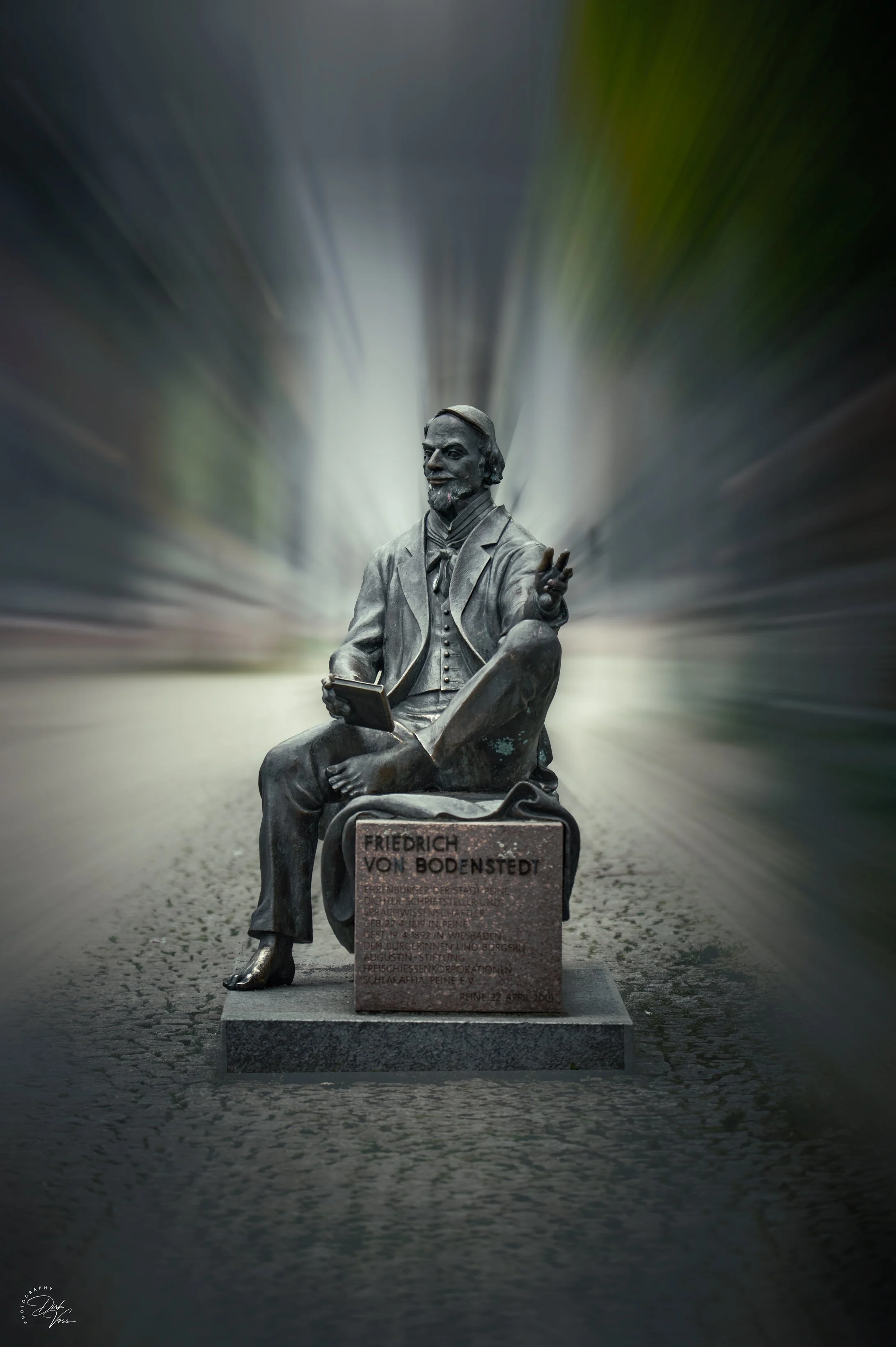 Bronzestatue eines sitzenden Mannes mit Bart, der ein Buch hält, auf einem alten Stadtplatz. Der Gedenkstein unten trägt die Inschrift: "Friedrich von Bodenstedt."