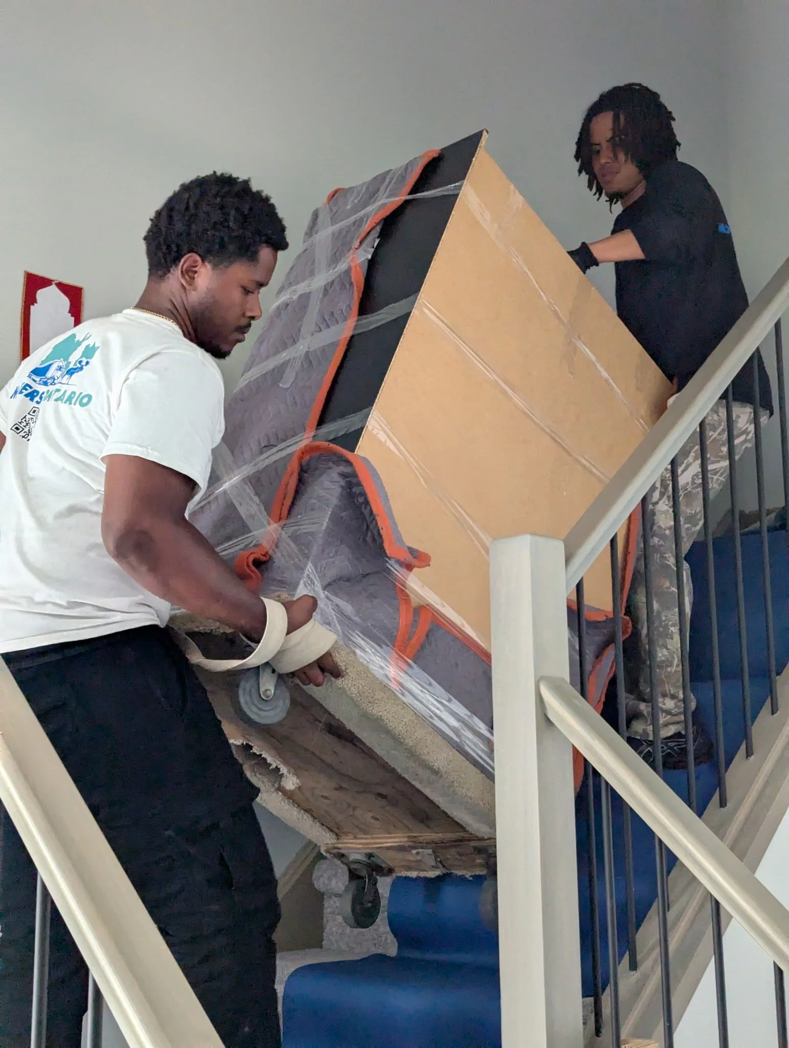Two workers are moving a large, wrapped piece of furniture or equipment up a staircase.