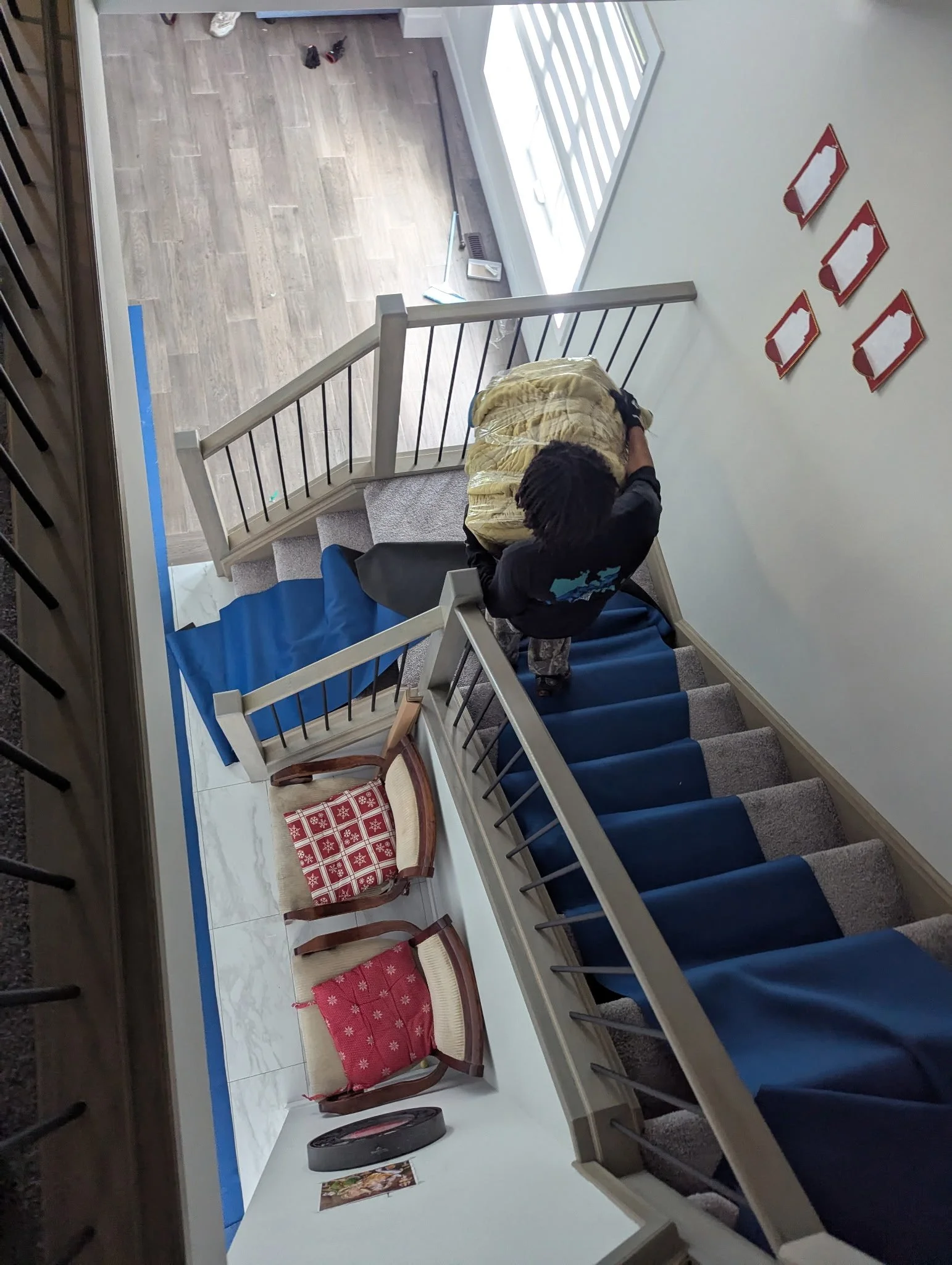 A person with dreadlocks, wearing a black shirt, is cleaning the stairs with a yellow cloth. The stairs have blue carpet runners, and the railing is white. There are two chairs with red and white cushions at the bottom of the stairs. On the wall to the right, there are four red framed decorations. The flooring at the top of the stairs is light wood, transitioning to marble at the bottom.