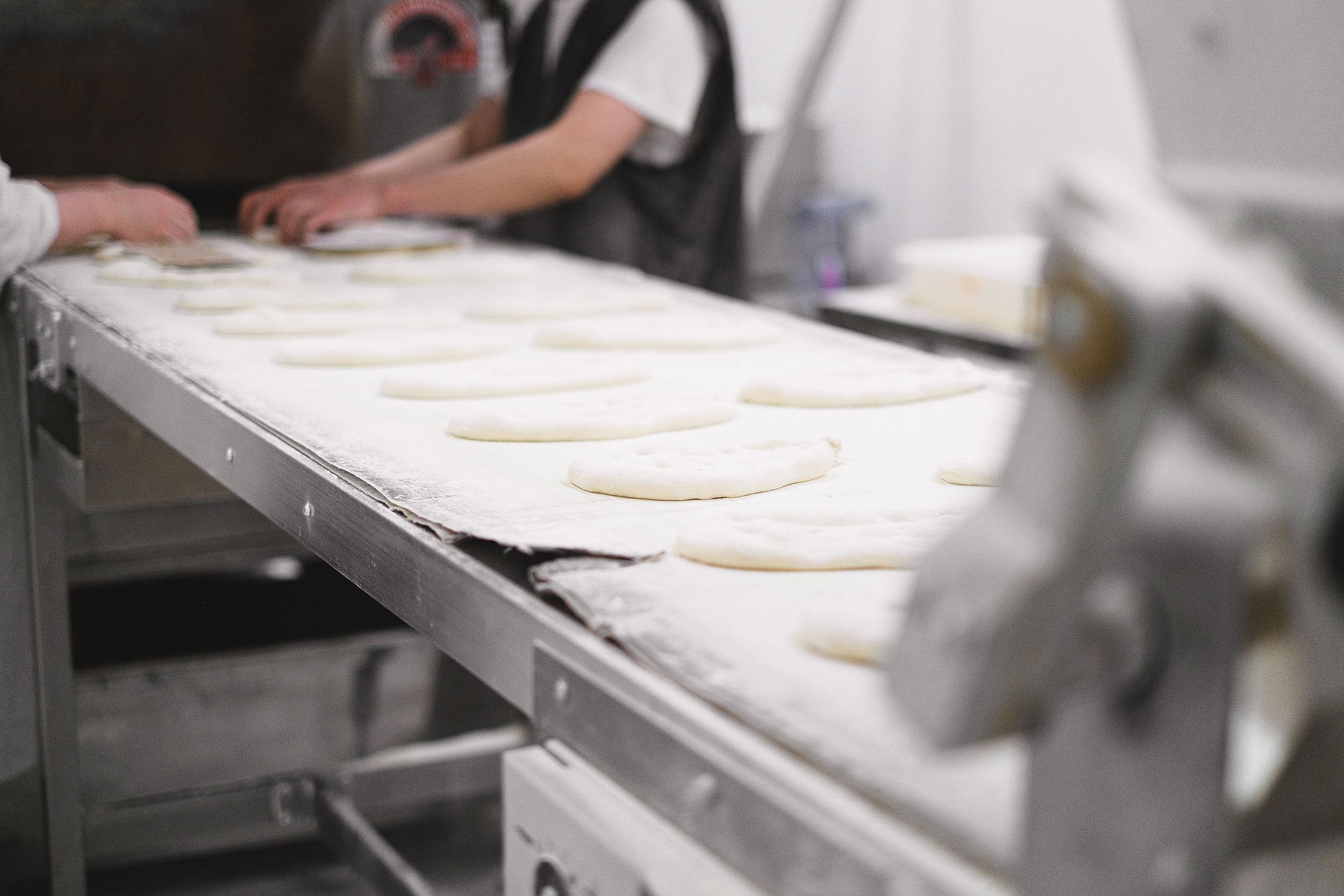 Rows of hand-shaped dough portions on a floured conveyor belt in a bakery production line, with staff shaping more dough in the background.