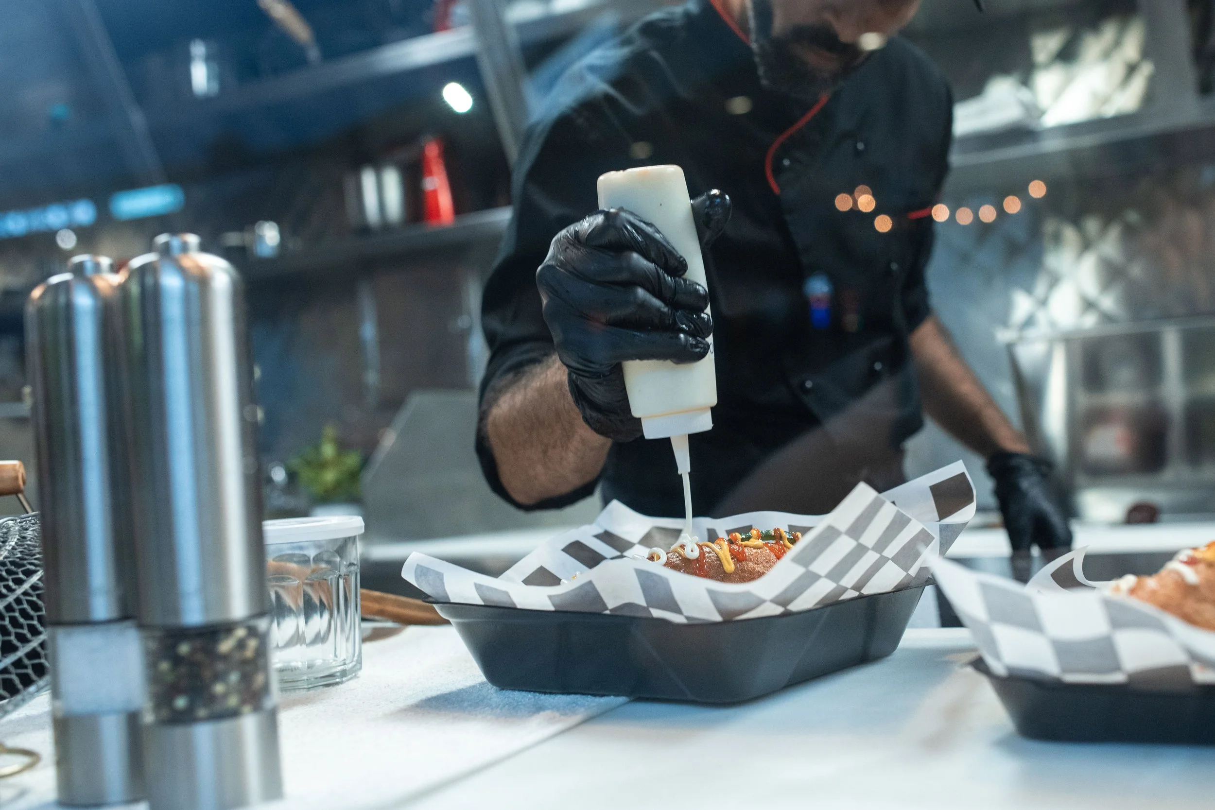 Chef preparing food in a cloud kitchen, drizzling sauce over a hot dog for a delivery order