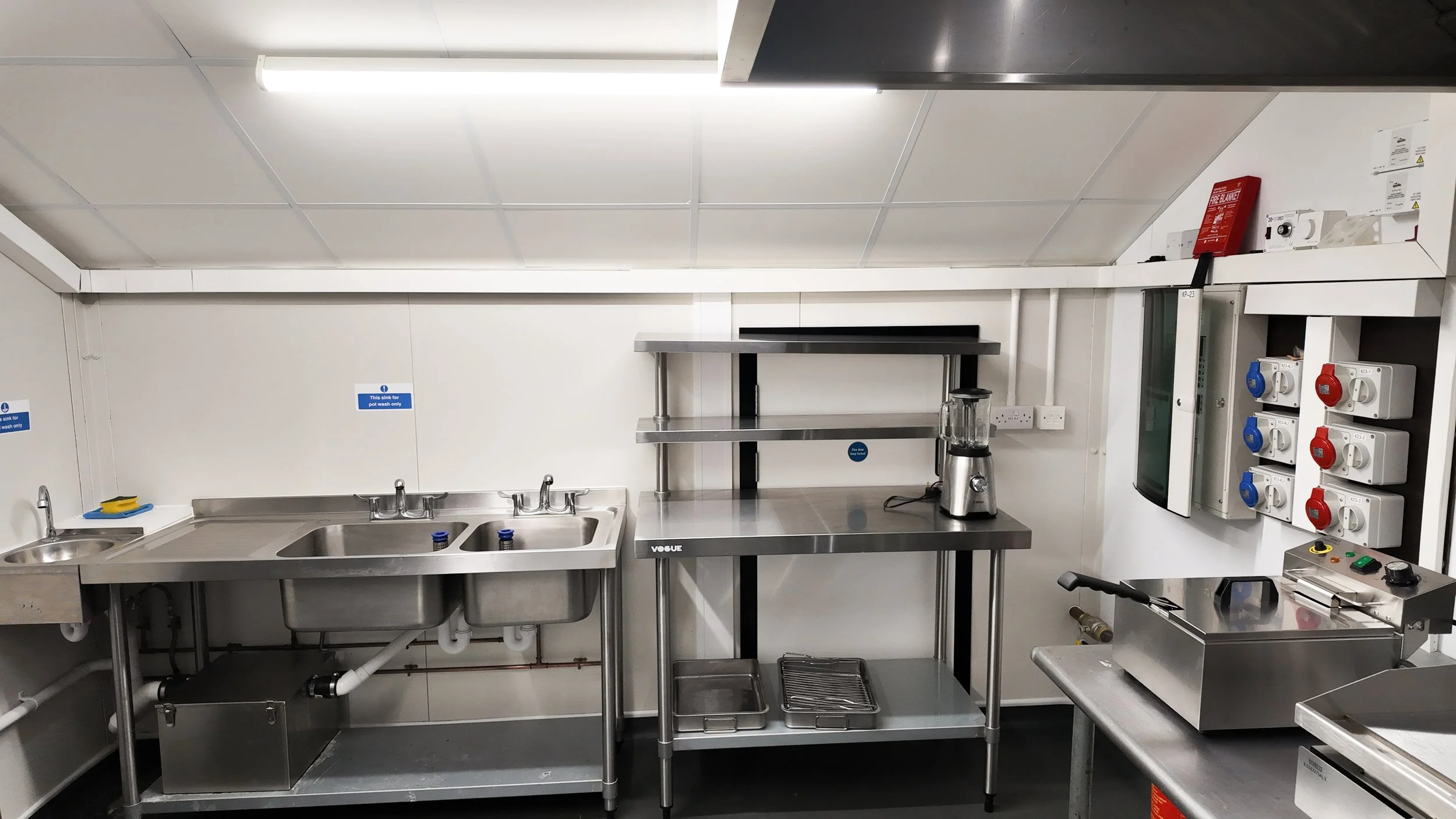 Fully equipped commercial kitchen with a stainless steel double sink, hand-wash basin, prep table with overshelf, countertop fryer, extraction canopy and three-phase power sockets, ready for short-term hire.