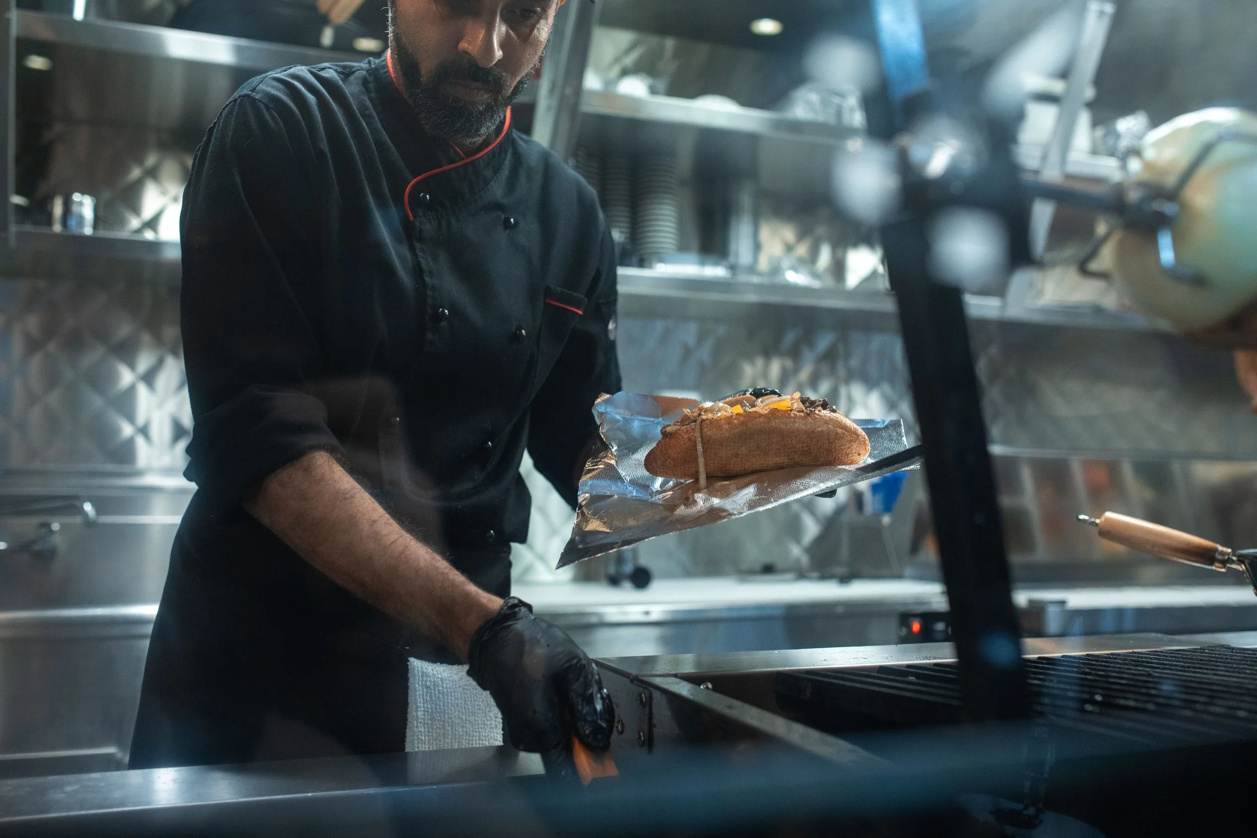 Chef in a commercial kitchen lifting a filled sub roll on foil from a grill, with tongs and stainless steel equipment in the foreground.