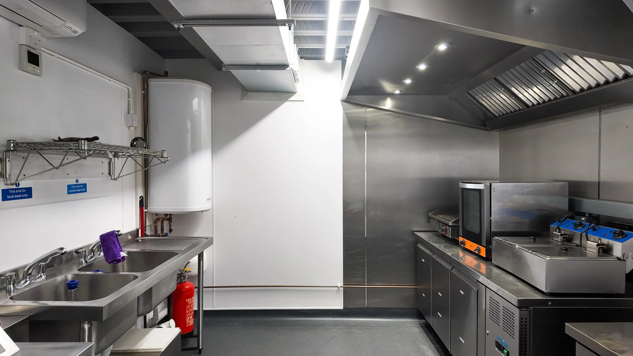 Commercial kitchen with stainless steel cladding, extraction canopy, convection oven, griddle, twin countertop fryers, under-counter fridges and a double sink for food and utensil washing.
