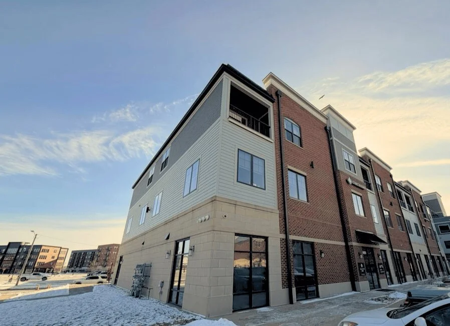 Exterior view of a multi-story residential building with brick and siding facades, large windows, balconies, and a sidewalk with snow. Other apartment buildings are visible in the background during sunset.