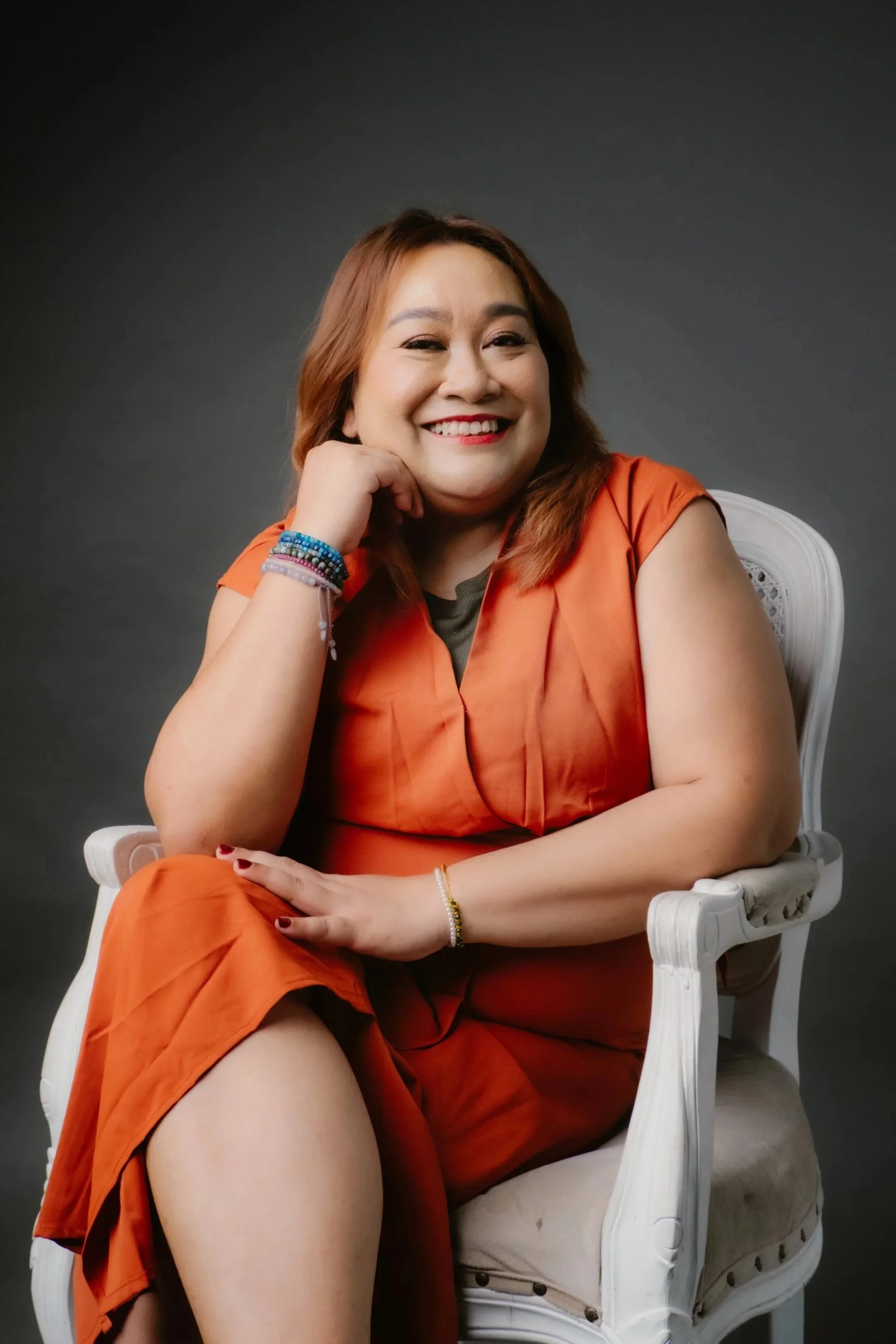 A woman with medium-length light brown hair, smiling and sitting in a white chair, wearing an orange sleeveless dress and colorful bracelets, against a dark gray background.