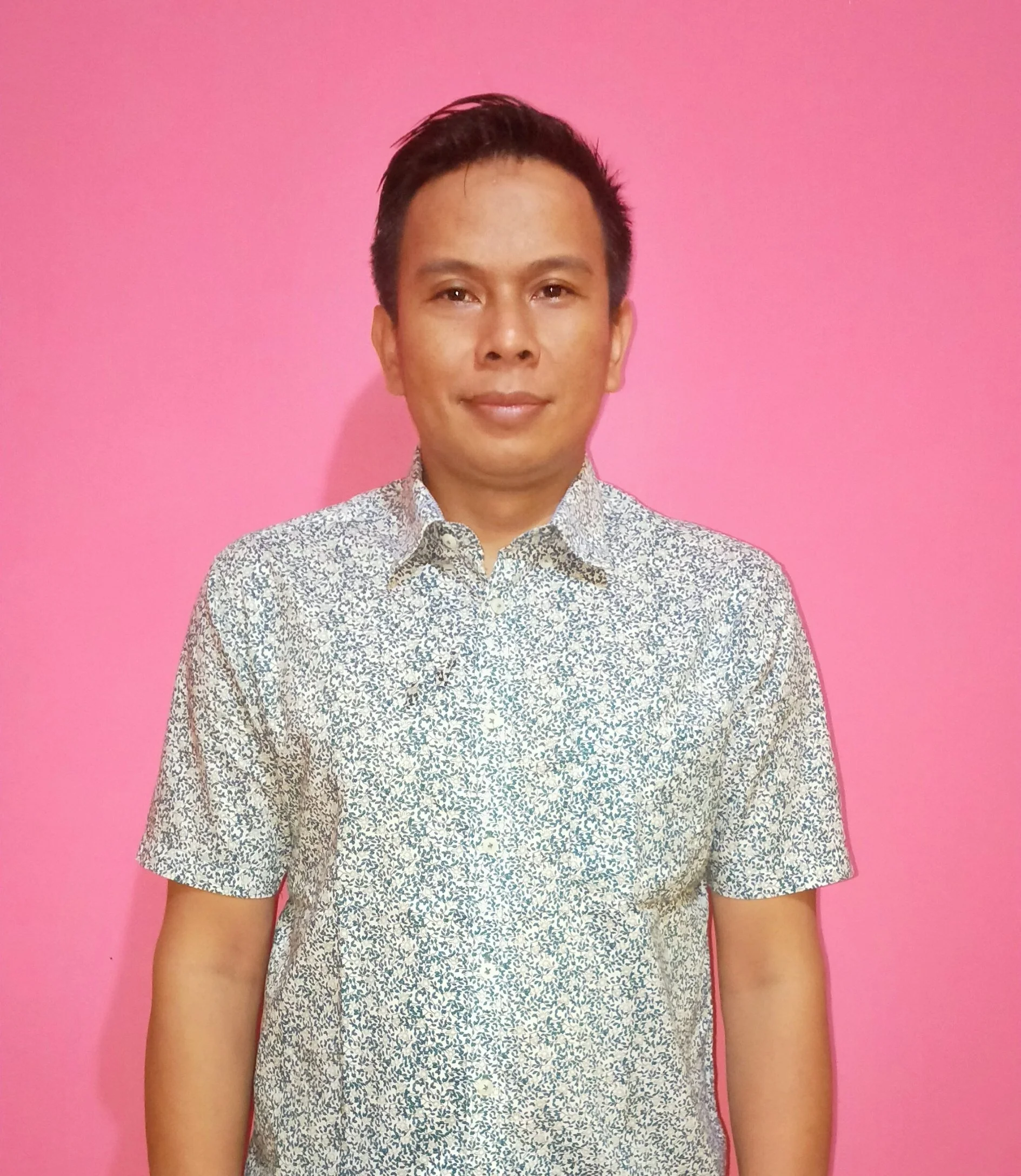 Portrait of a man in a patterned shirt standing against a pink wall.