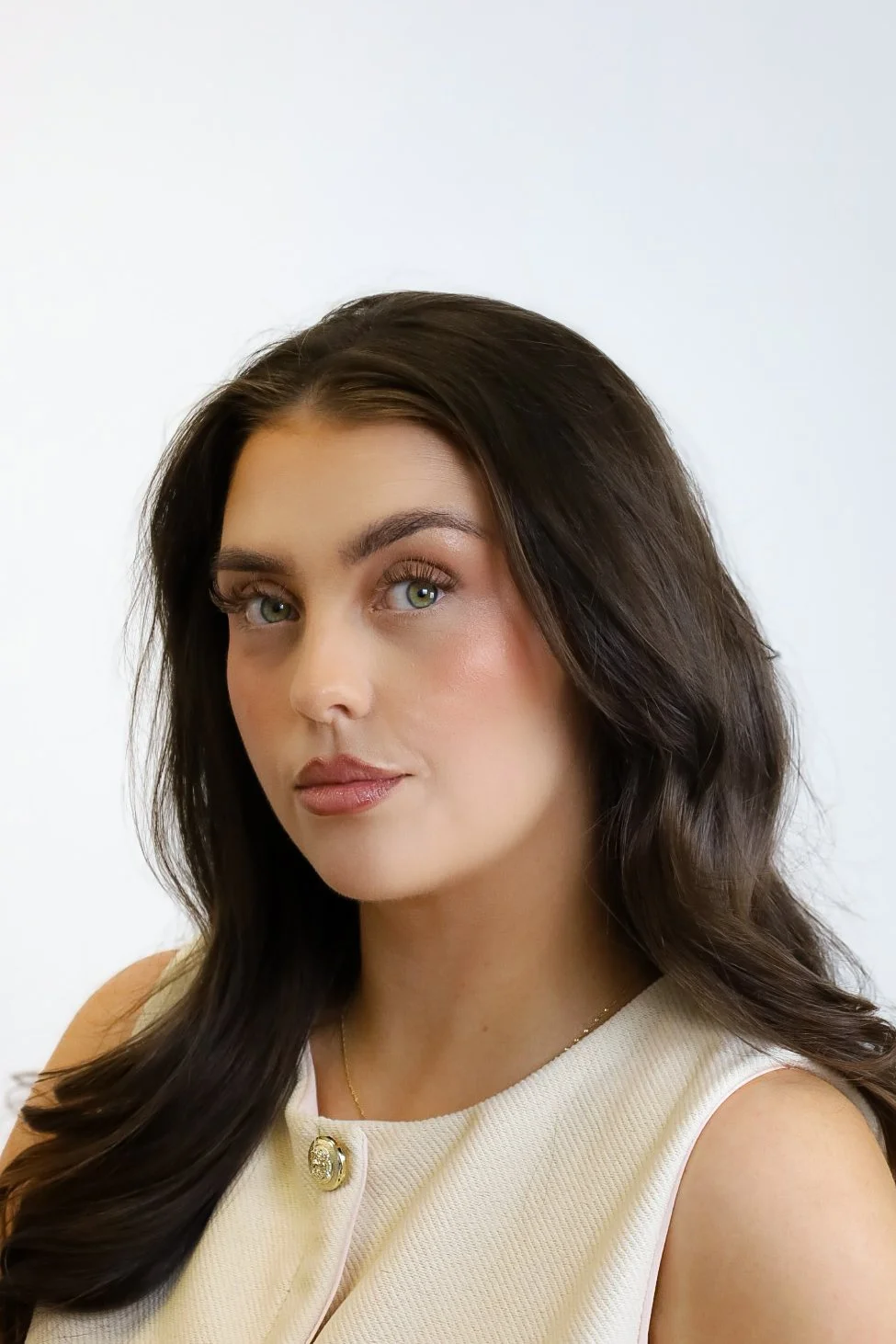 A woman with long brown hair, light skin, and green eyes, wearing a black top, standing indoors with beige vertical blinds in the background.