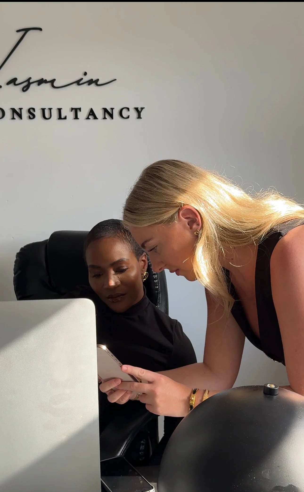 Two women are looking at a smartphone together in an office setting, with a computer screen and a sign that reads 'GEORGIA JASMIN.  CONSULTANCY' in the background.