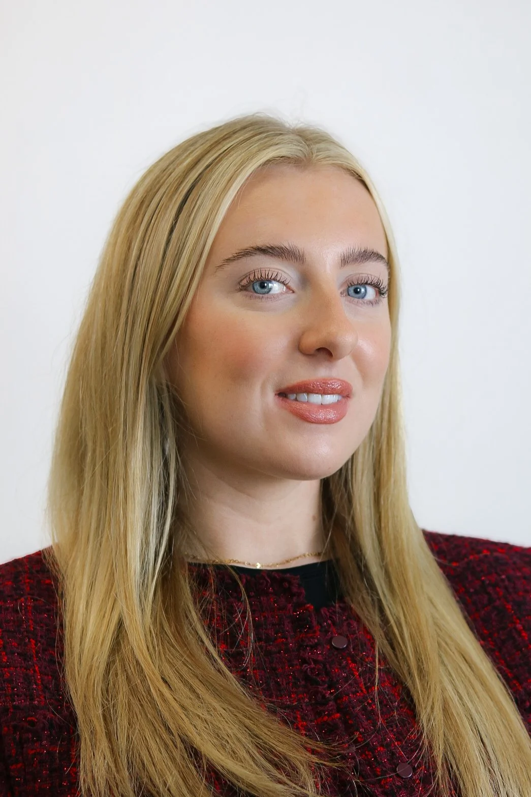 A portrait of a woman with blonde hair, blue eyes, wearing a black top, earrings, and lipstick, standing in front of a background with white curtains.