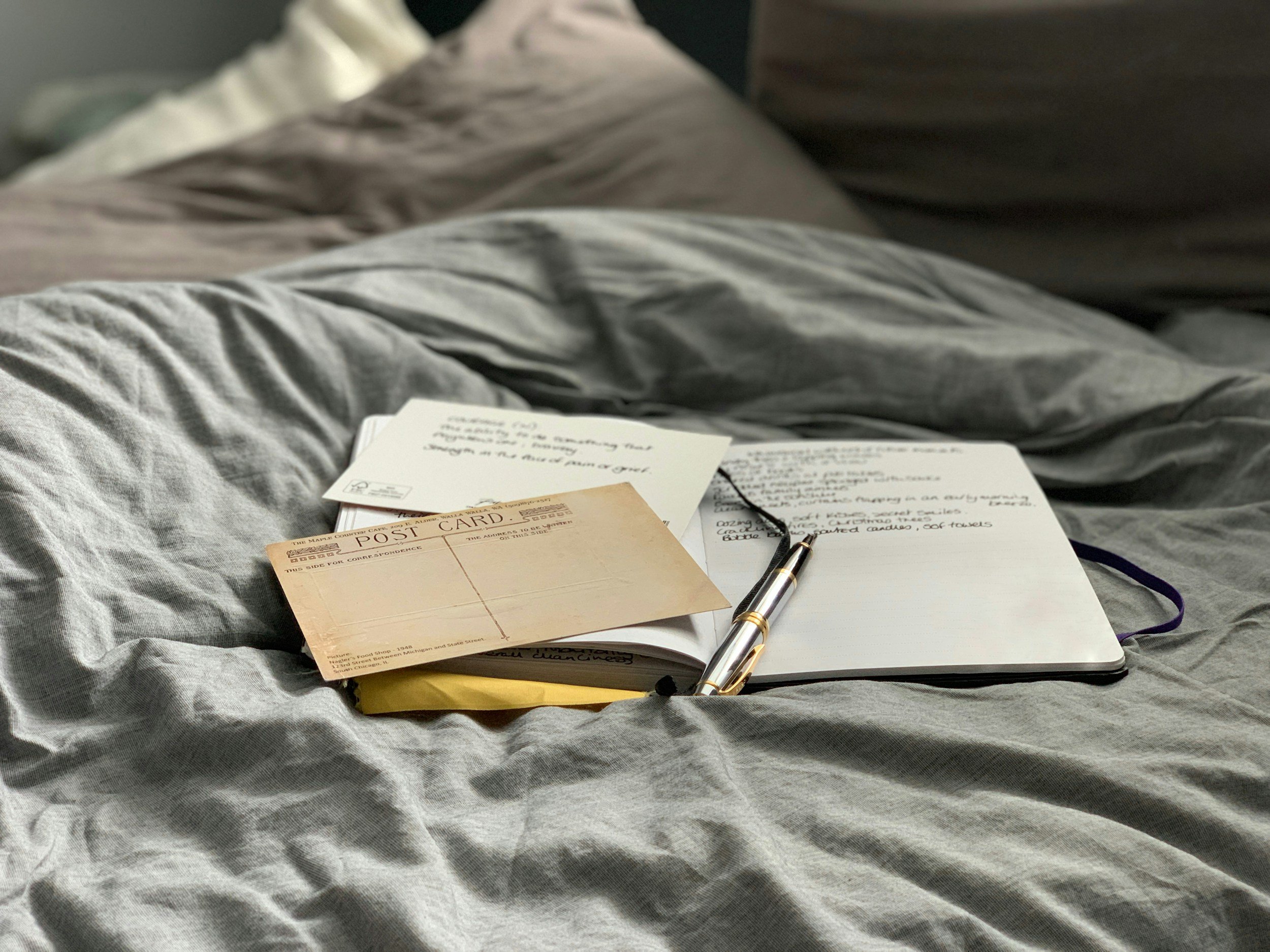 Open notebook, postcard, handwritten notes, and a pen resting on a bed with gray sheets.