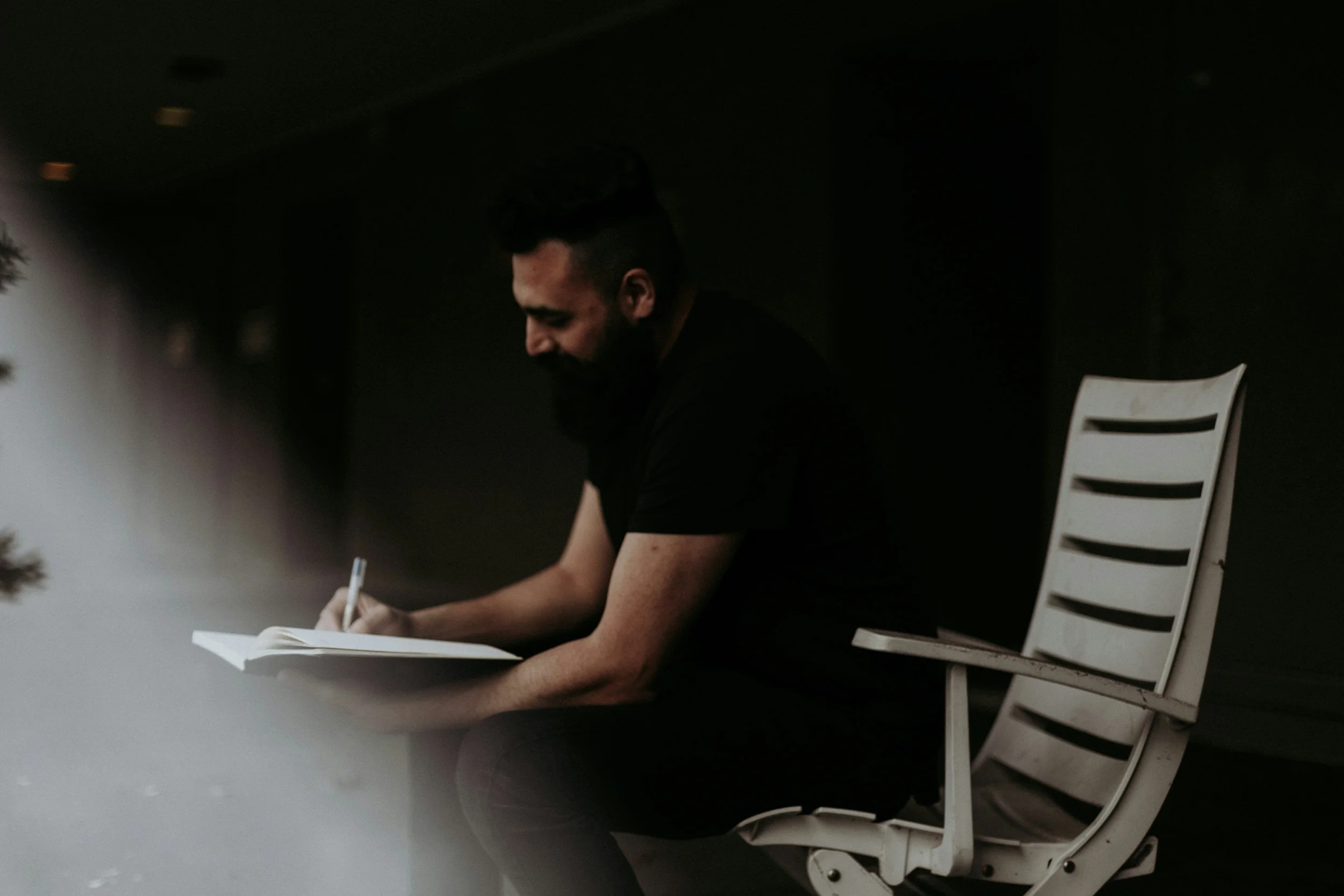 A man with a beard sitting in a white chair, writing in a notebook with a pen, in a dimly lit room.