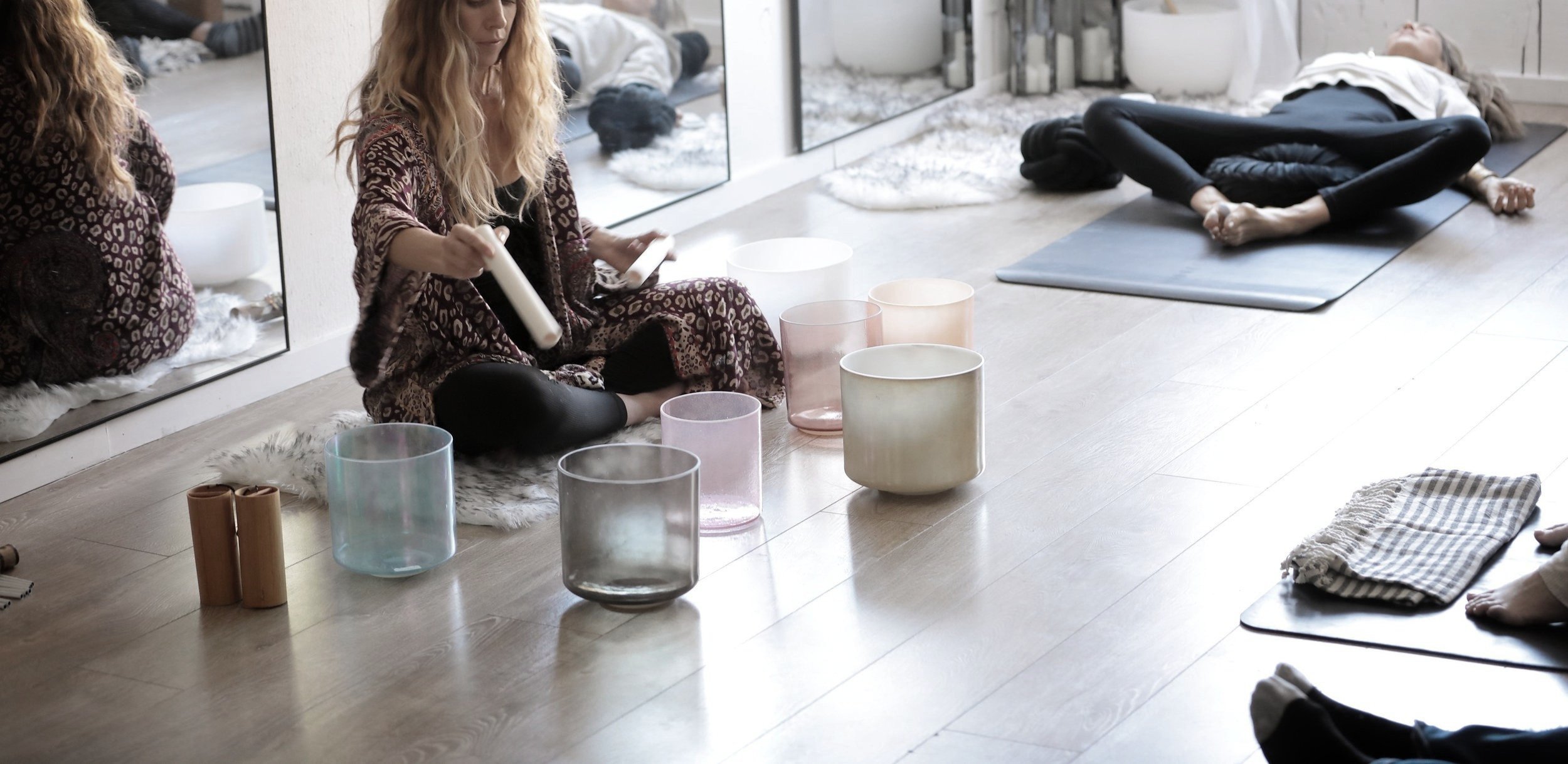 People engaging in a crystal singing bowl meditation session in a studio, with various bowls on the floor and participants lying on mats.