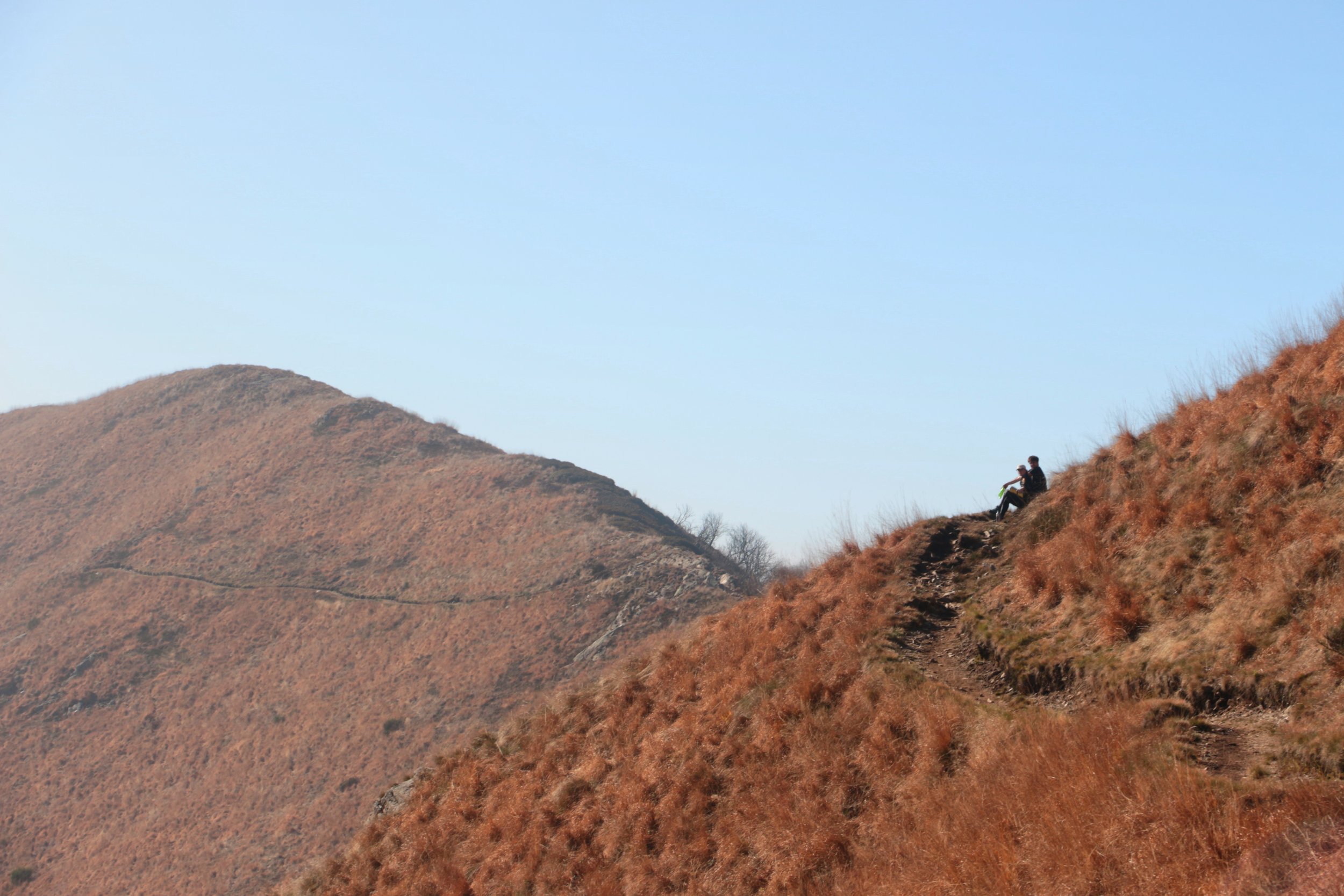 Two people sitting on a grassy hillside, looking at the view, with a mountain in the background under a clear blue sky.