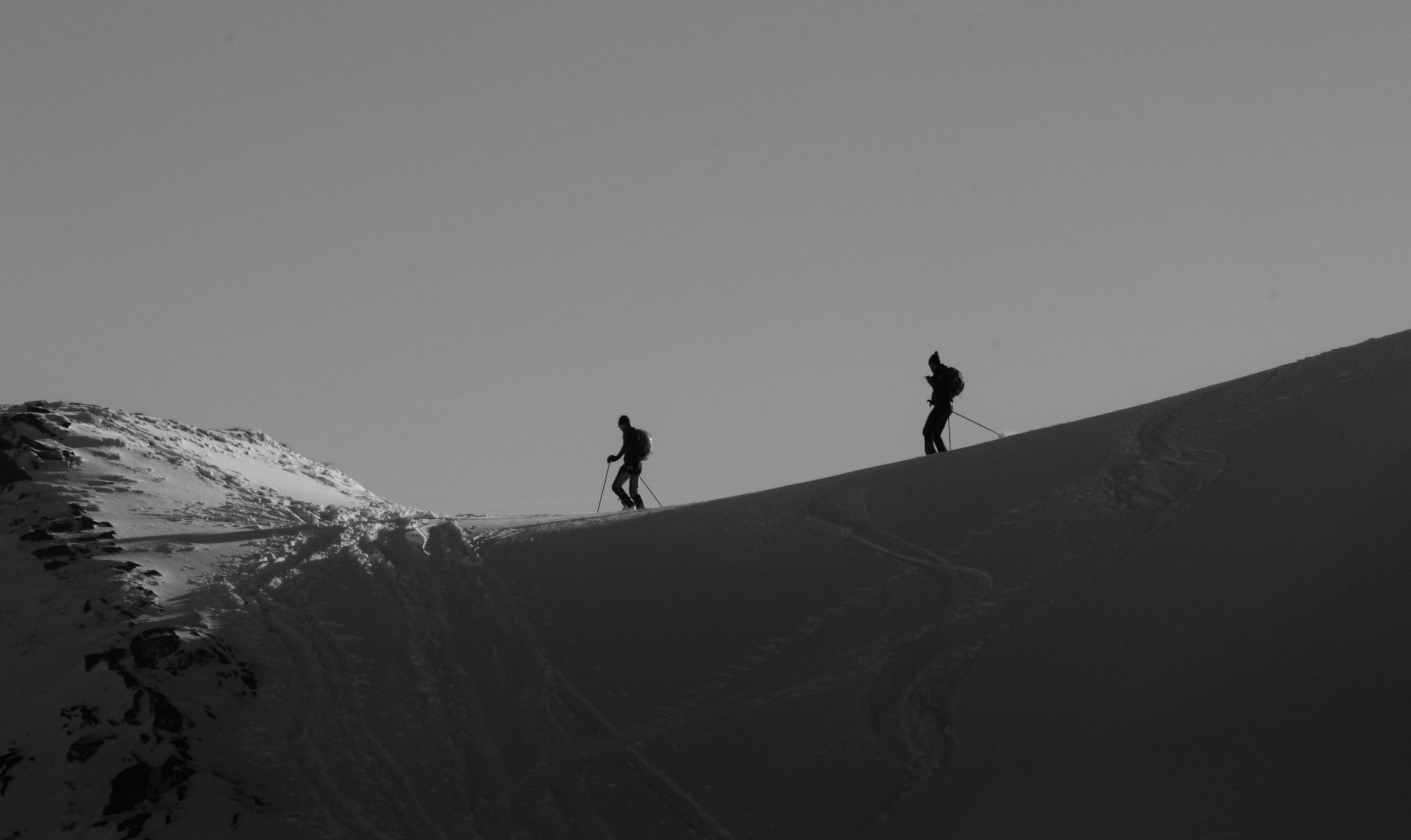 Two skiers climbing a snowy mountain slope at sunset or sunrise, with visible ski tracks.
