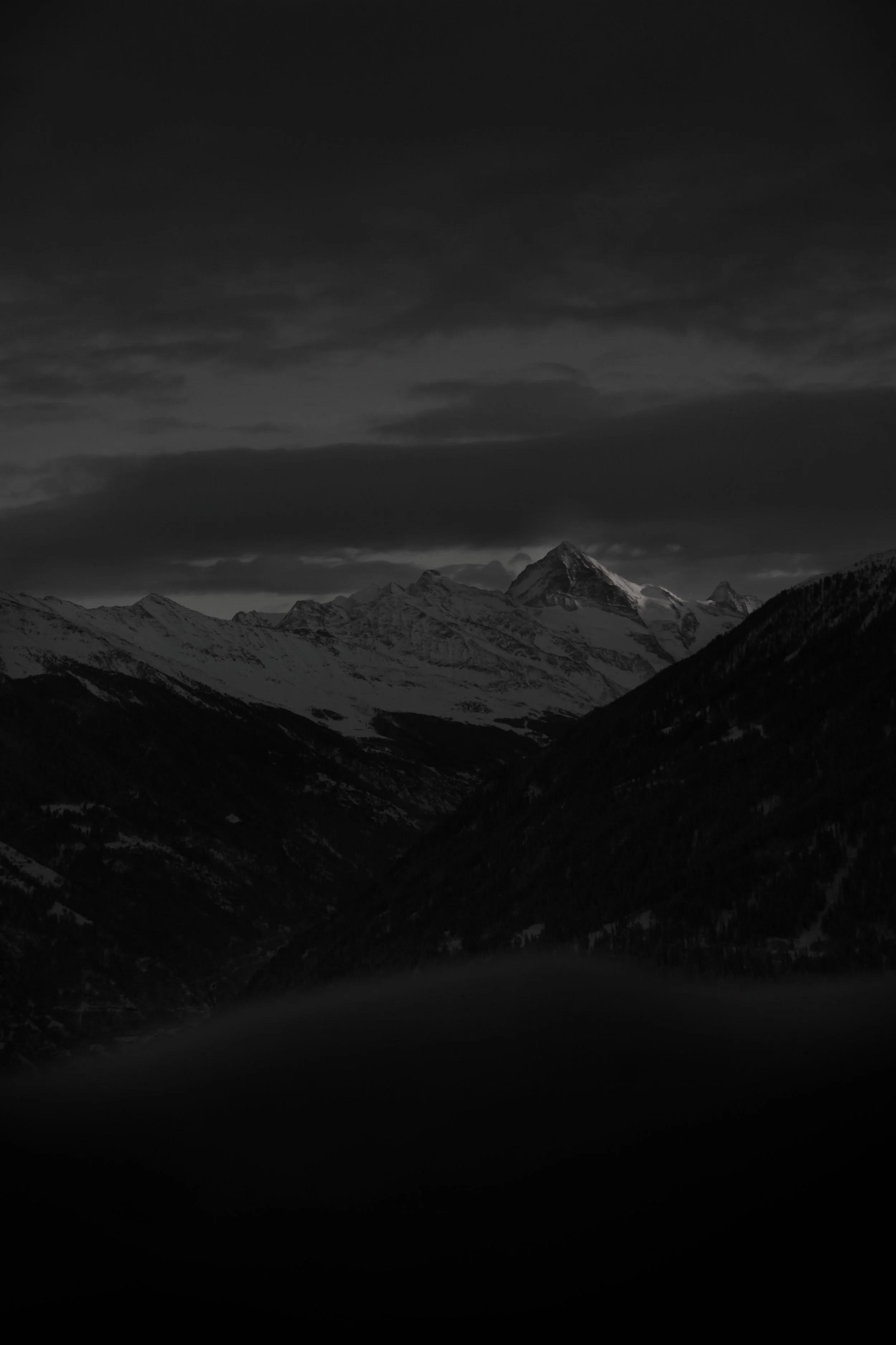 Black and white photo of snow-covered mountain peaks under a cloudy sky.