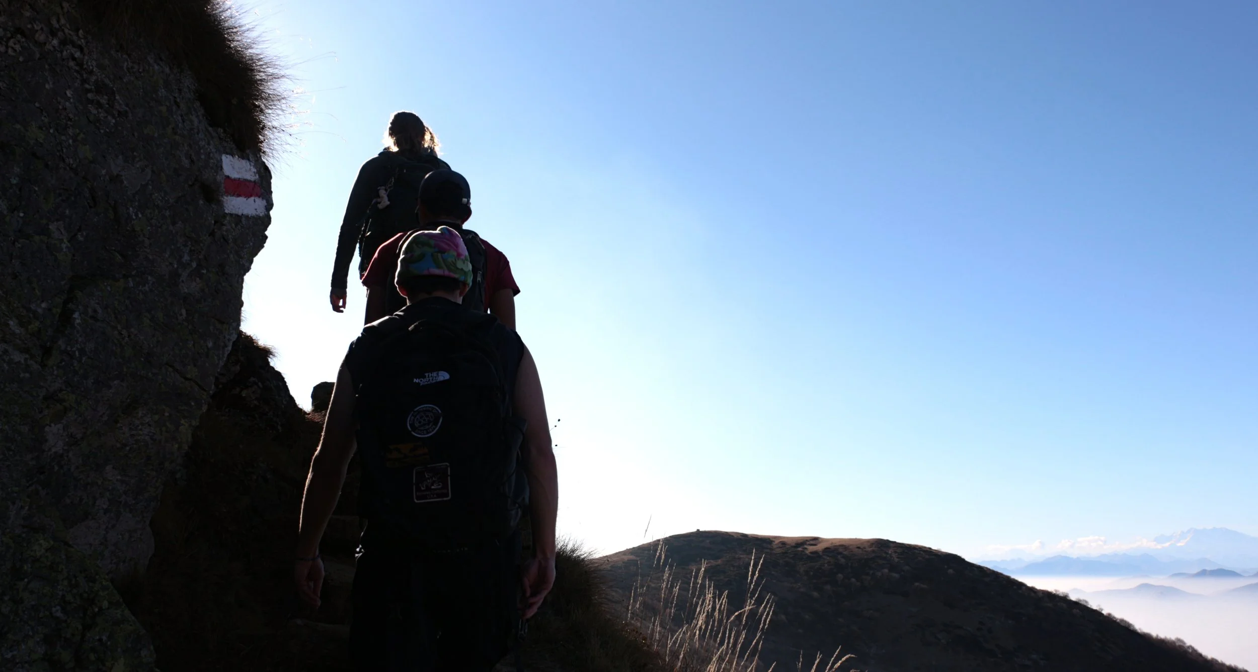 Group of hikers walking along a mountain trail during daytime with clear blue sky and distant mountain range in the background.