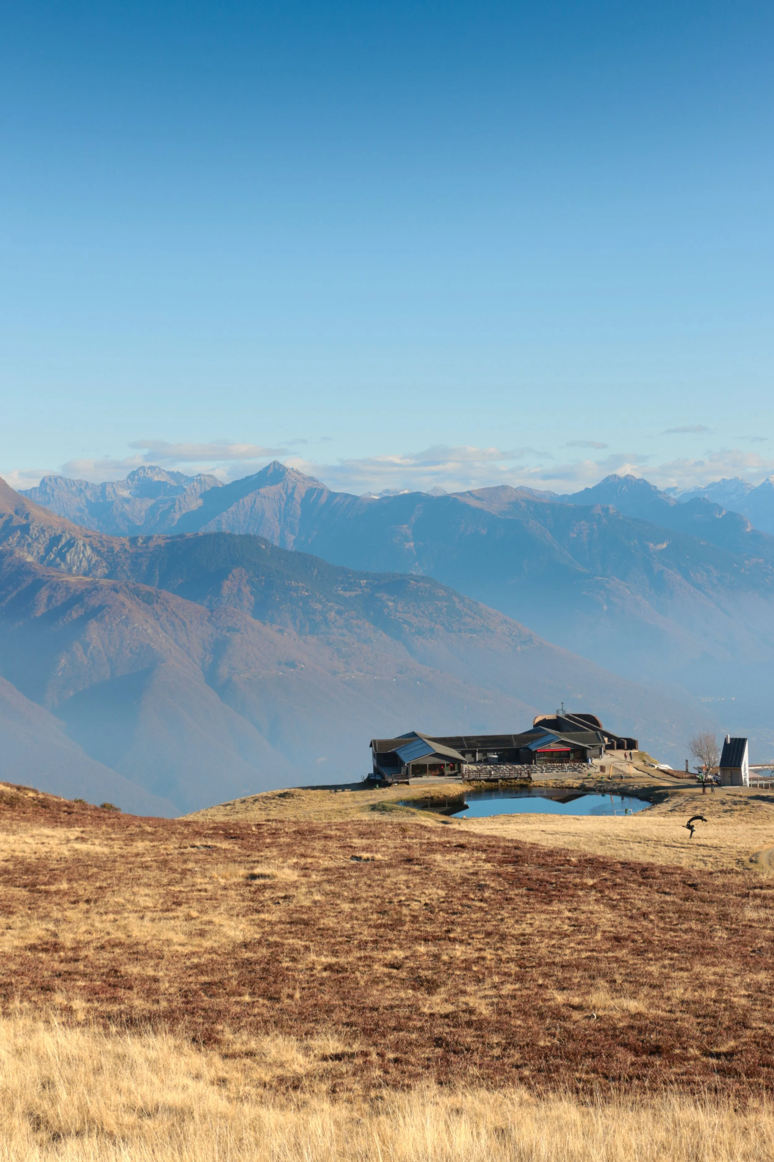 A mountain landscape with a building and small pond in foreground, and tall mountains in the distance under a blue sky.