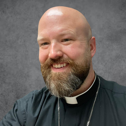 Headshot of a smiling male priest with a beard and bald head, wearing a black clerical shirt with a white collar against a gray background.