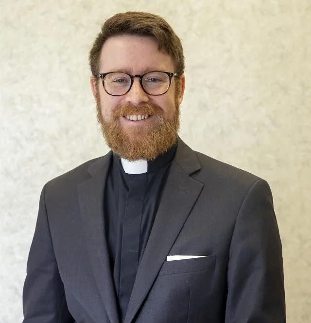 A smiling man wearing glasses and a clerical collar in a dark suit, standing against a plain background.