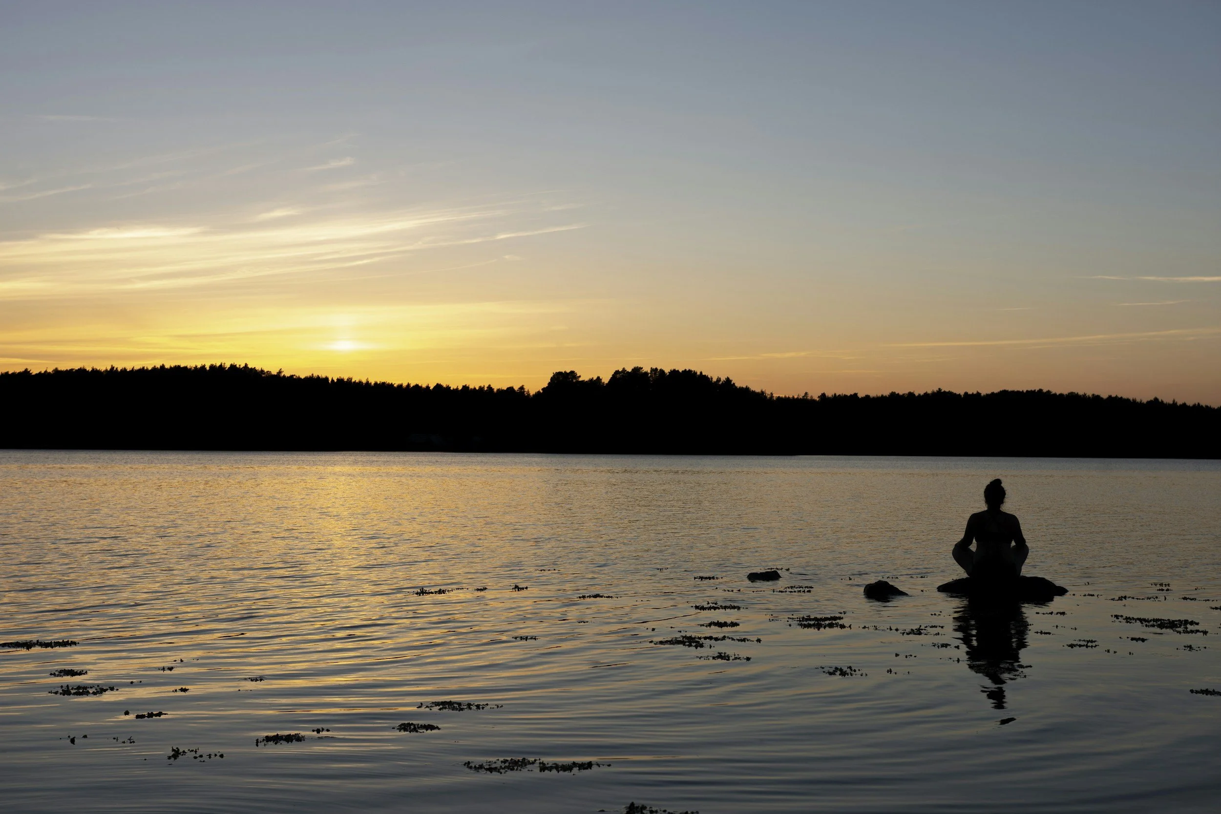 A person practicing yoga by a lake at sunset, sitting cross-legged on a rock with a scenic view of the water, trees, and a partly cloudy sky.