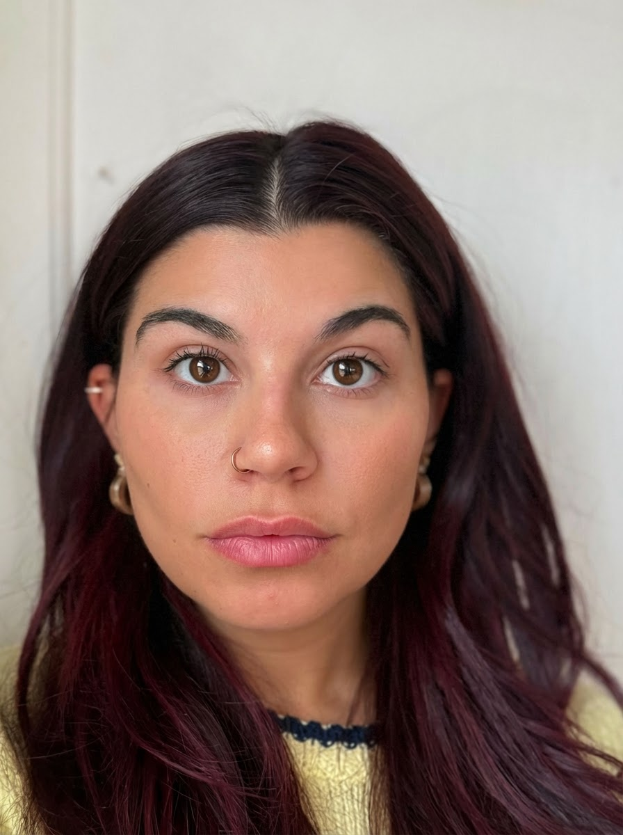 A woman with dark brown and purple hair, wearing a nose ring, earrings, and a yellow top, looking directly at the camera.