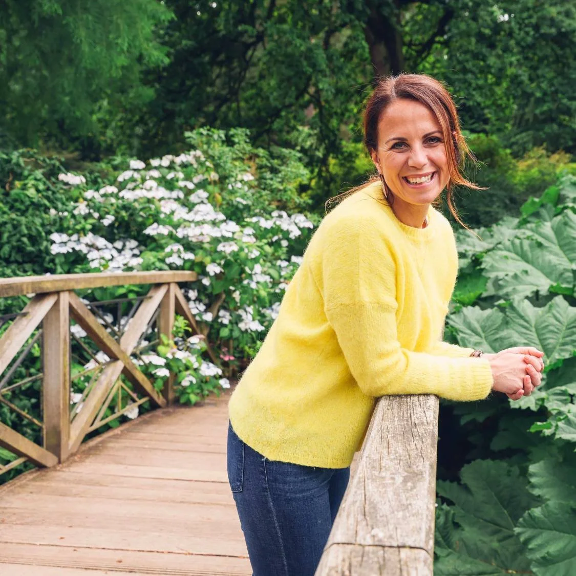 Woman in yellow sweater leaning on wooden bridge in lush garden with white flowers, smiling.