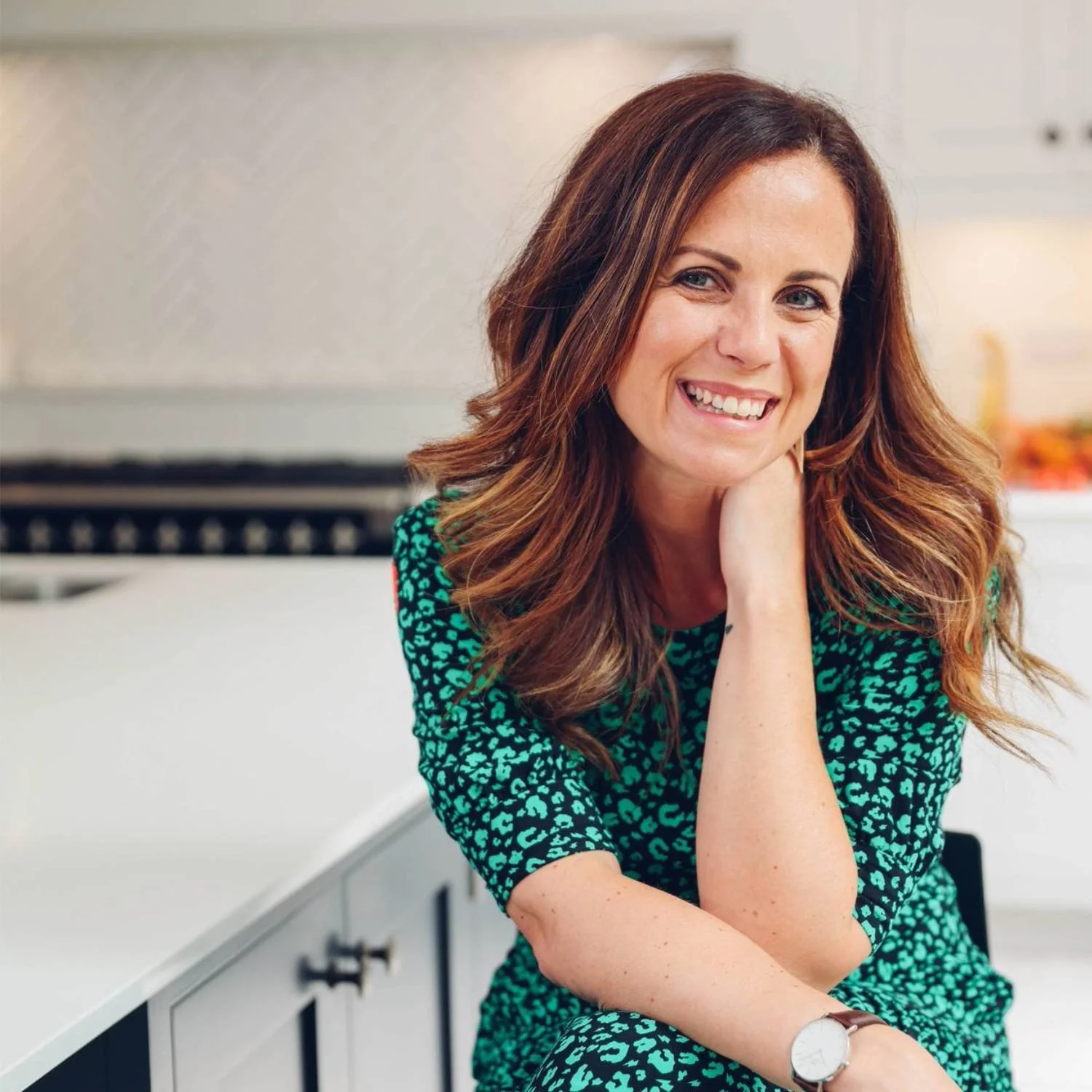 Smiling woman sitting in a kitchen wearing a green patterned dress.