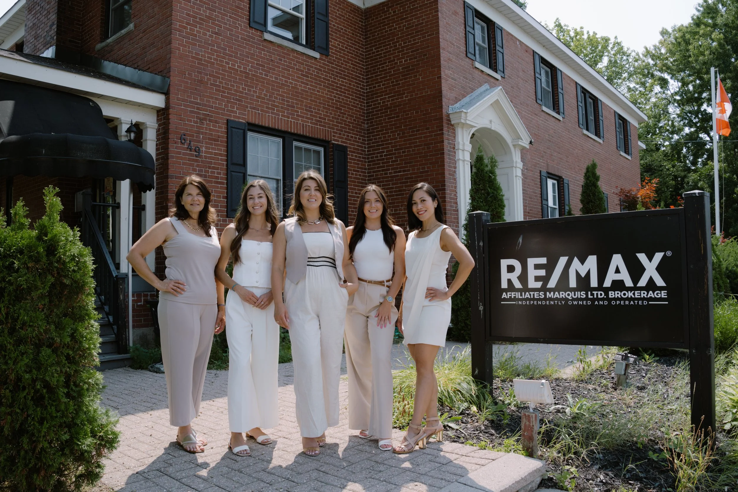 Five women dressed in white standing in front of a brick building with a RE/MAX sign in a landscaped area.