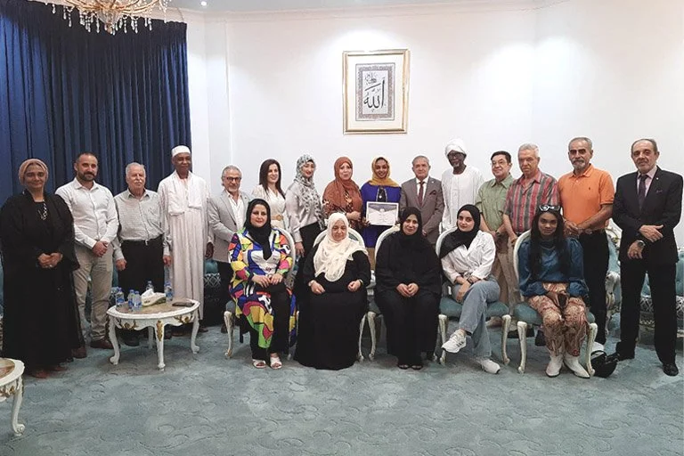 A group of people posing indoors in a formal setting, with some seated and others standing. They are dressed in a mix of traditional and modern attire. The setting includes elegant furniture, a framed artwork on the wall, and a chandelier.