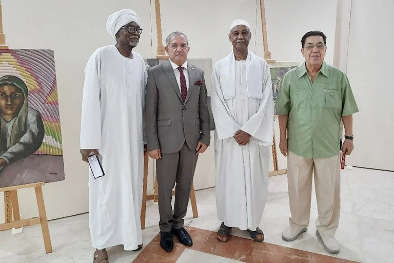 Four men standing in front of artwork on easels, featuring portraits. Two men are wearing traditional white attire, one is in a suit, and one is in casual green and beige clothing.