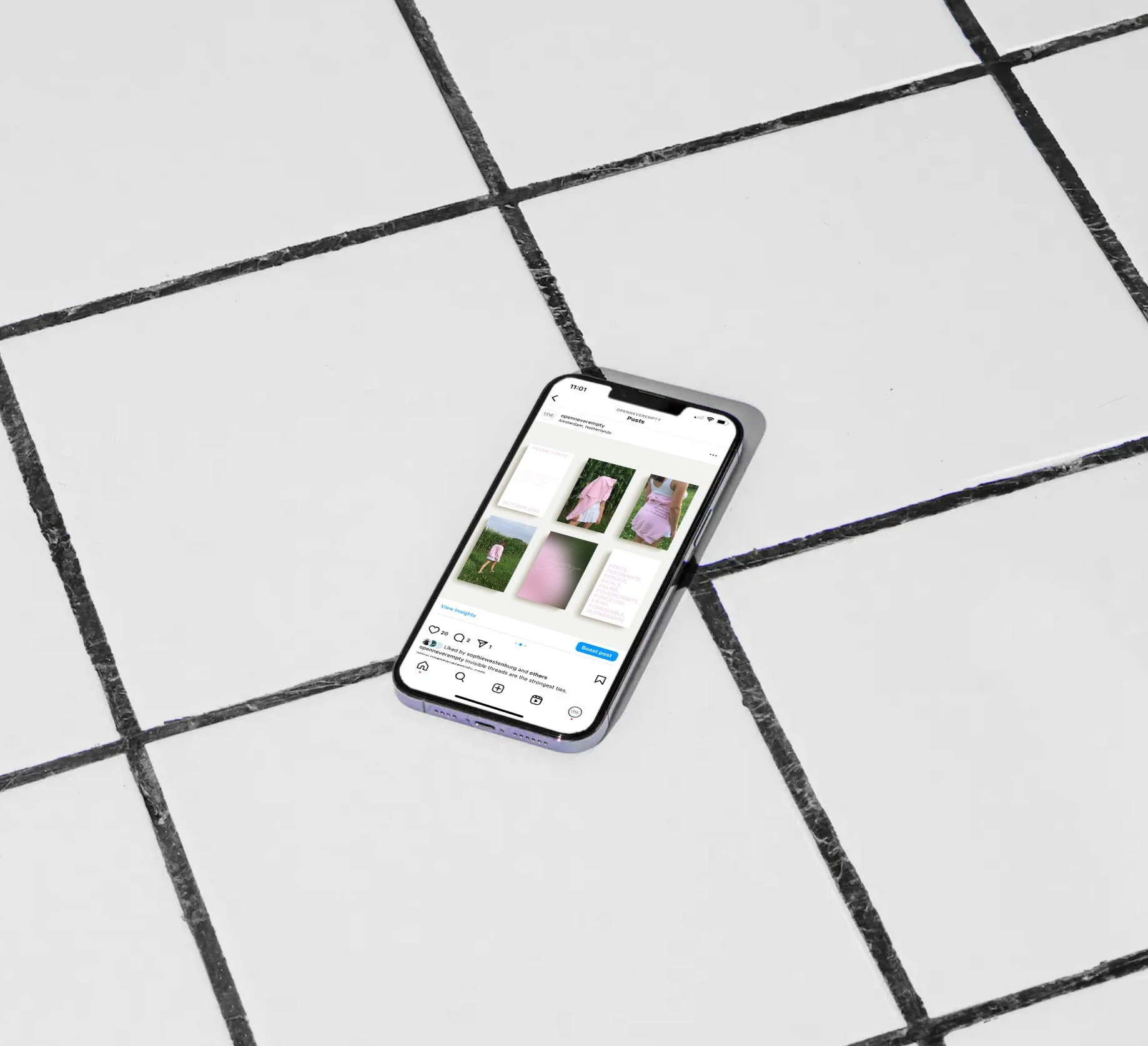 A smartphone lying on a tiled floor displaying an Instagram profile with images of a pink dress and a person in a pink dress outdoors.
