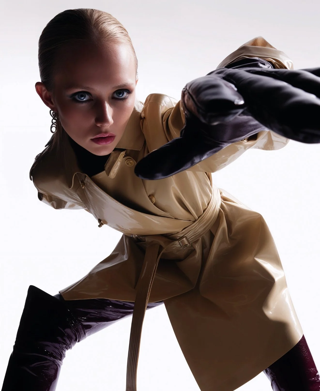 A woman in a tan trench coat and black gloves reaches towards the camera with a serious expression, against a plain white background.