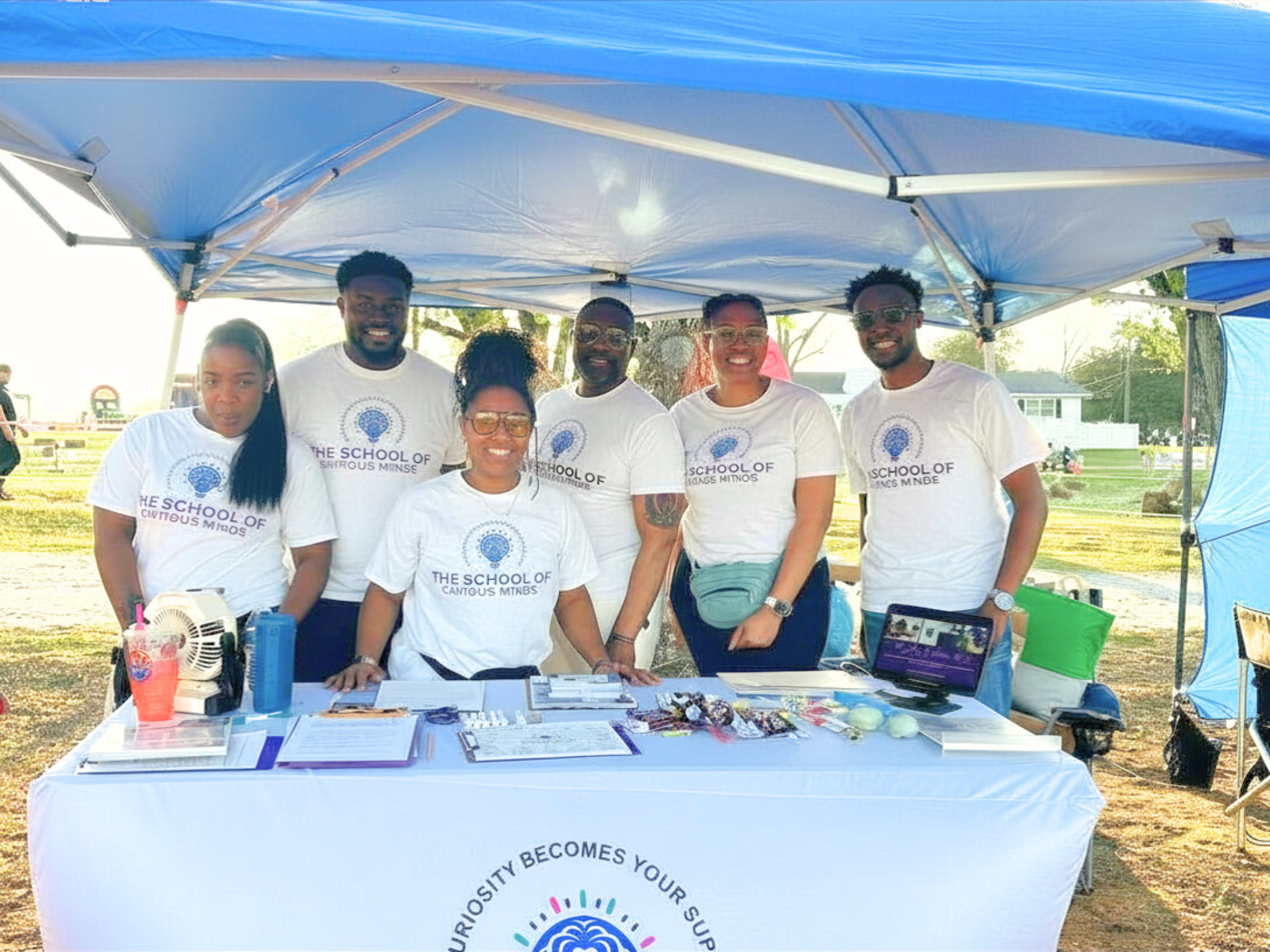 Group of six young adults standing behind a table under a blue canopy, wearing white T-shirts with a logo, at an outdoor community event.