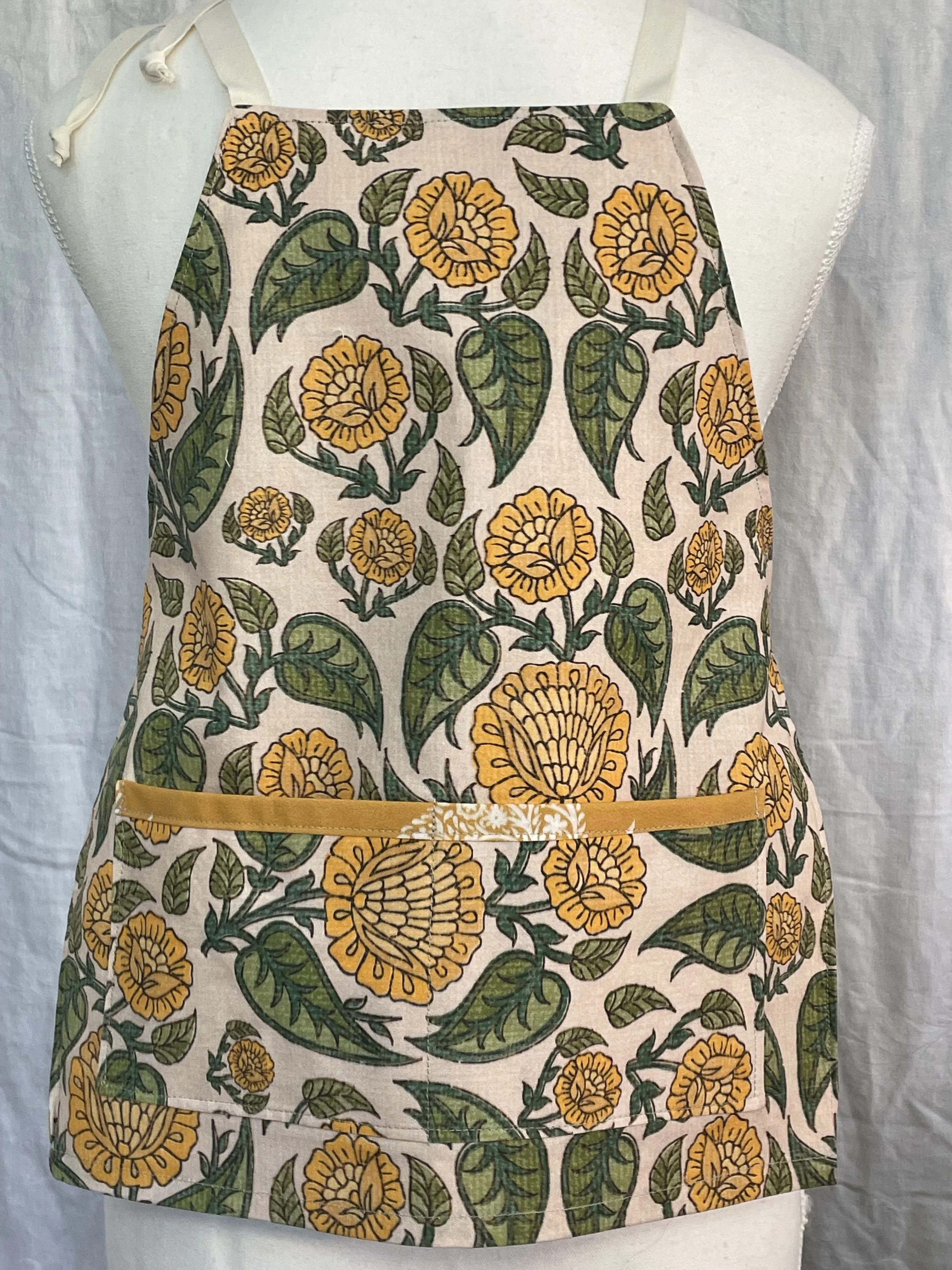 A child's apron with a yellow floral pattern that features large yellow flowers with green leaves. The apron has a yellow belt at the waist.