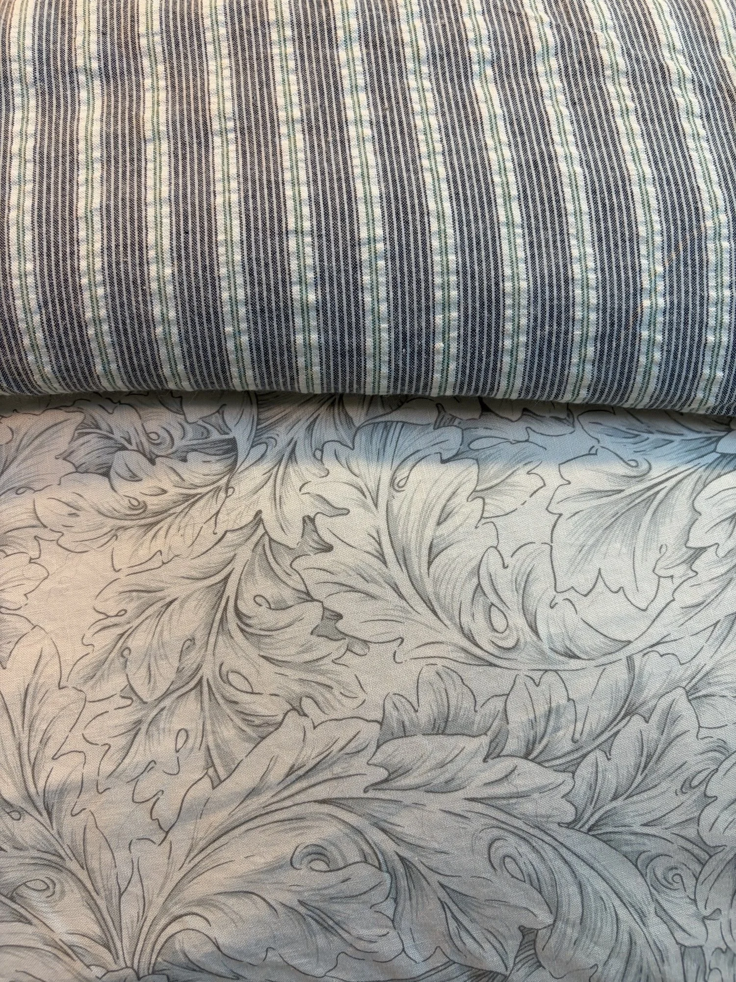 Close-up of layered fabrics featuring blue and white vertical stripes over a gray floral acanthus pattern.