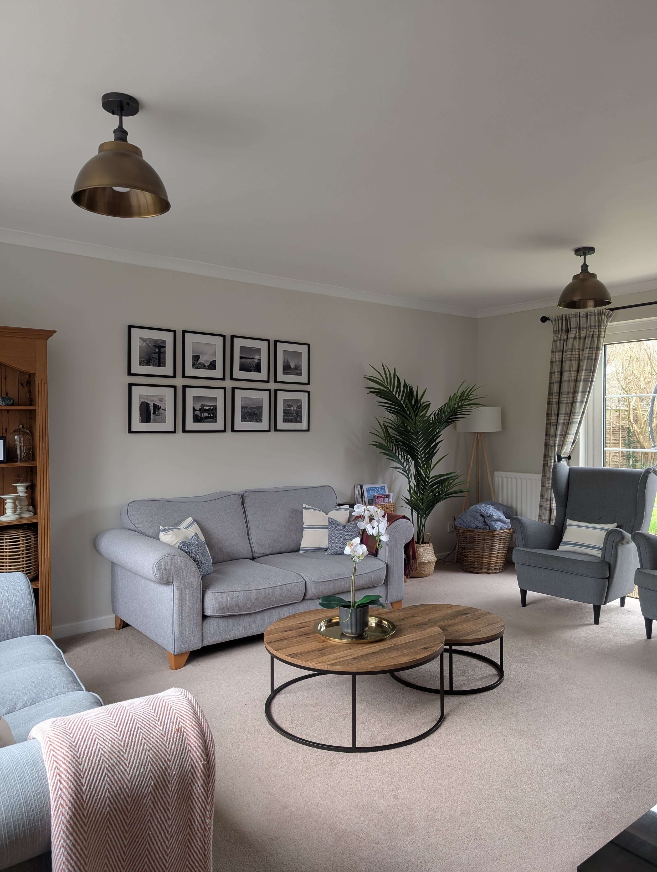 Living room with gray sofa, two matching armchairs, wooden coffee tables, framed black and white photos on wall, potted plant, floor lamp, window with plaid curtains, beige carpet.