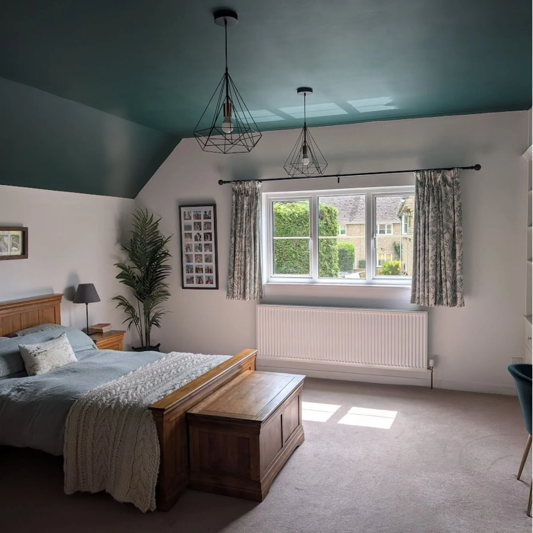 A bedroom with a window showing a view of neighboring houses and greenery. The room has a wooden bed with light-colored bedding, a blanket, pillows, and a side table. There is a tall plant and framed photos on the wall. Ceiling lights are hanging from the teal ceiling, which has a skylight. Curtains are drawn on either side of the window, and a radiator is below it.