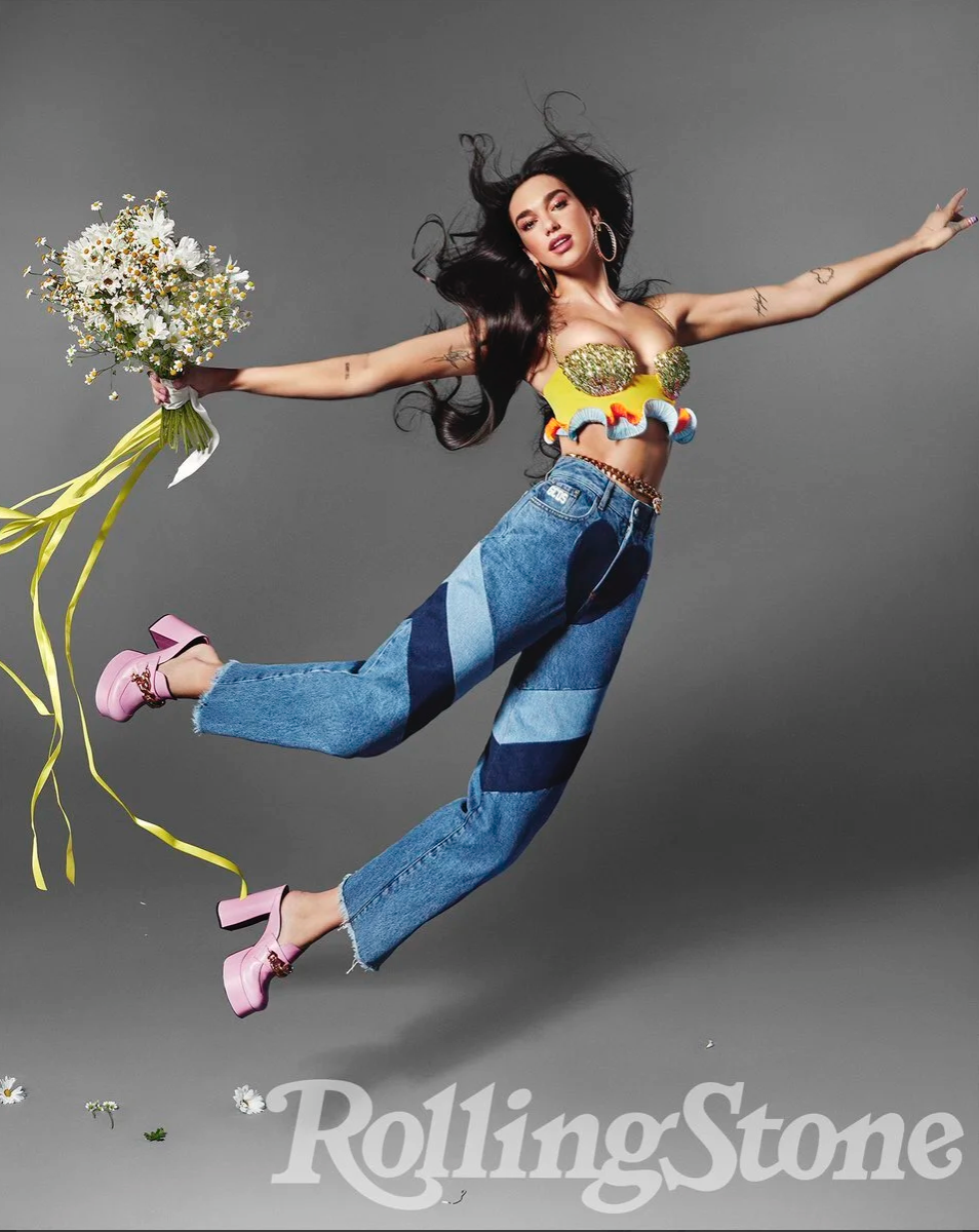A woman jumping with arms outstretched holding a bouquet of daisies. She has long dark hair, wearing hoop earrings, a colorful strapless top, baggy jeans, and pink platform shoes. The background is gray, with daisies and yellow ribbons floating around, and a Rolling Stone logo at the bottom.