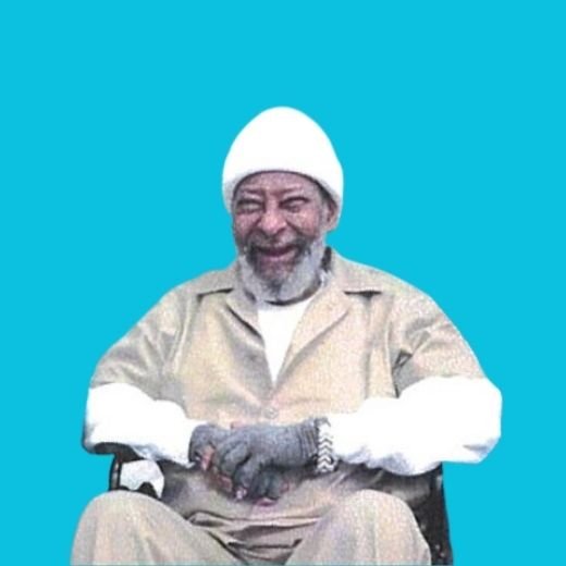 An elderly man with a gray beard and mustache, wearing a white beanie and beige clothing, smiling and sitting in a wheelchair against a bright blue background.