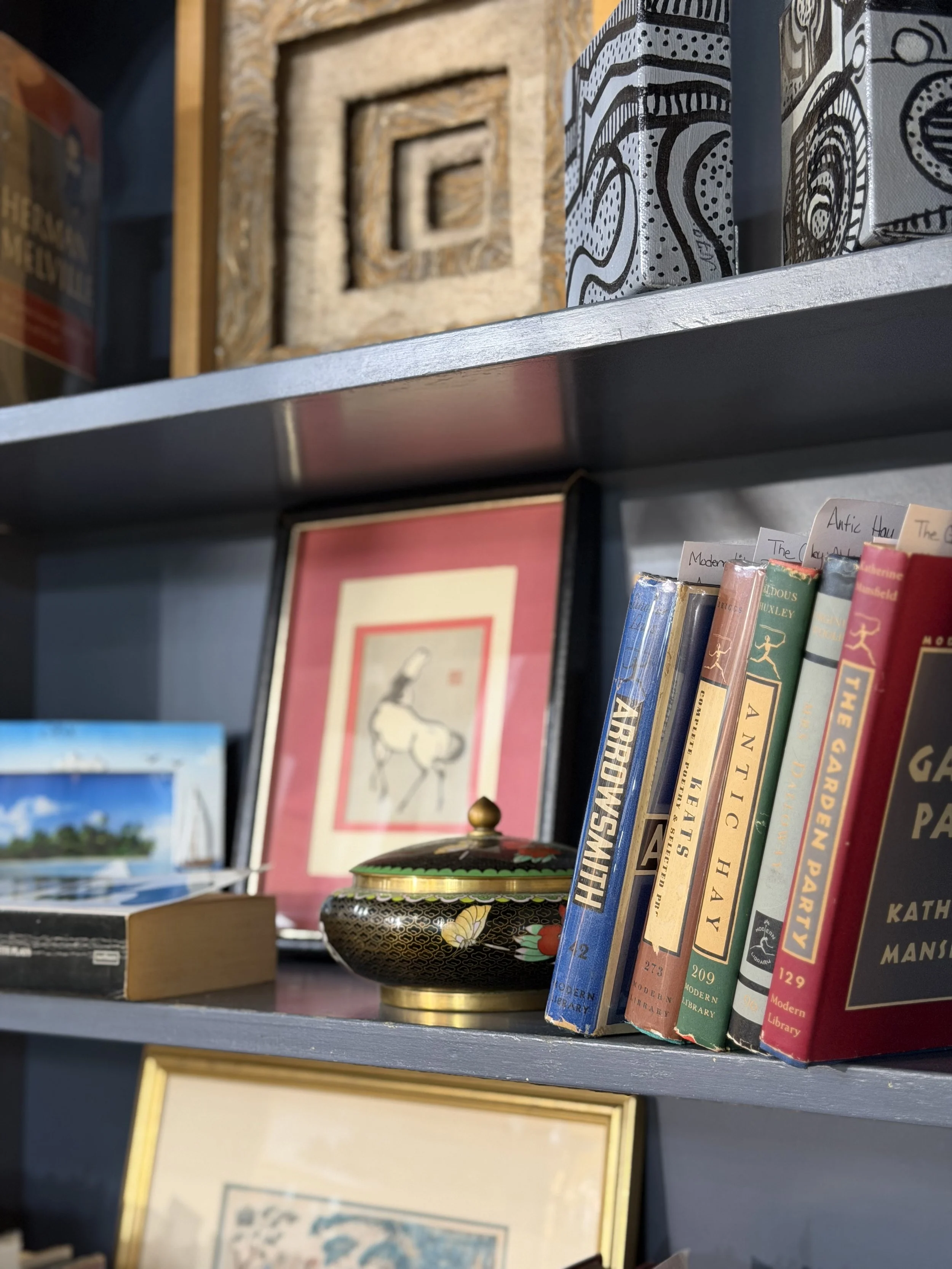 A bookshelf with various books, a framed abstract art piece, a decorative dish with a lid, and a landscape painting in the background.