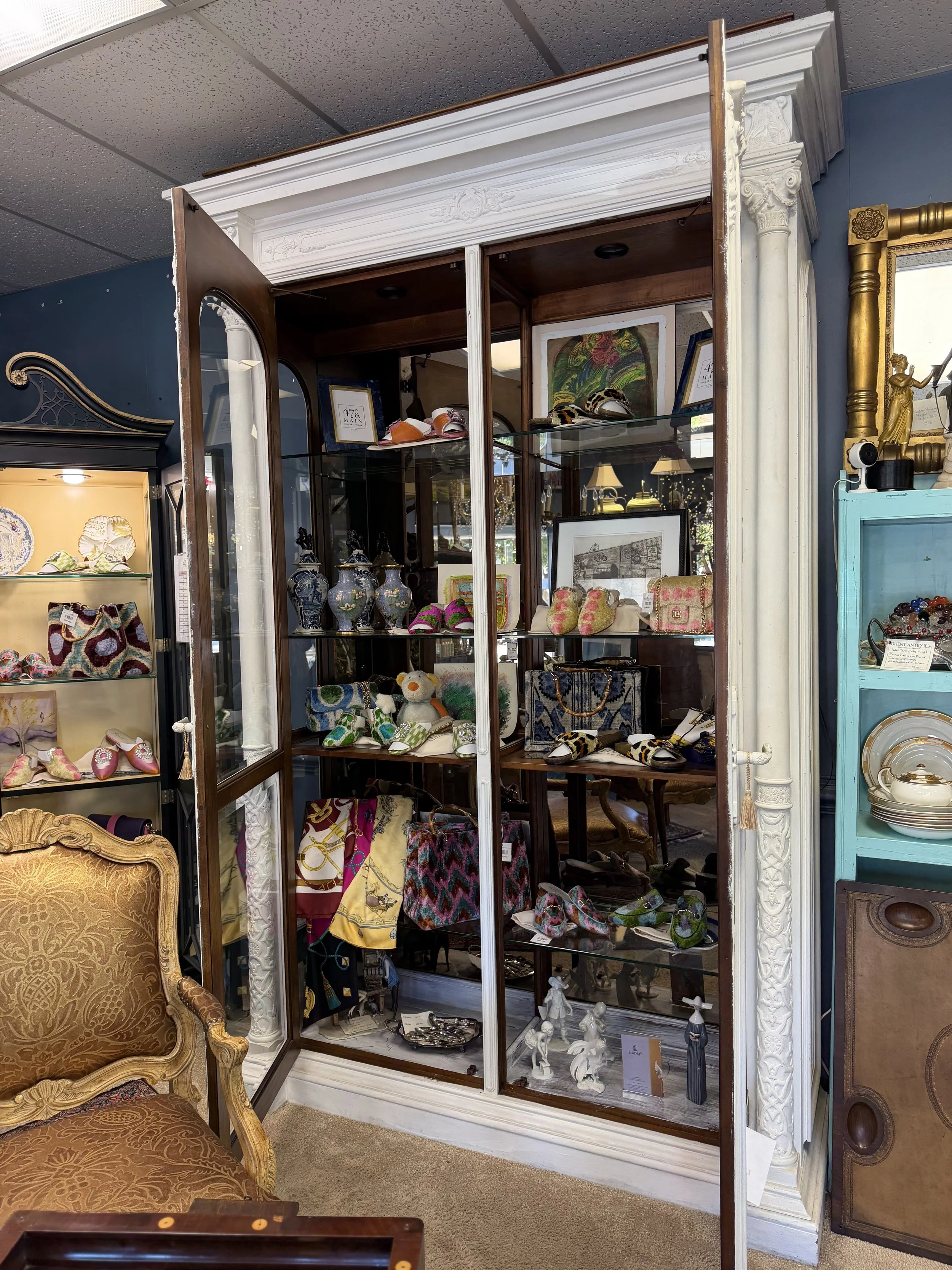A display cabinet filled with various decorative items, including pottery, plush toys, and scarves, is situated inside an antique store. To the left is a vintage armchair, and to the right are partial views of other shelving units with plates and oth