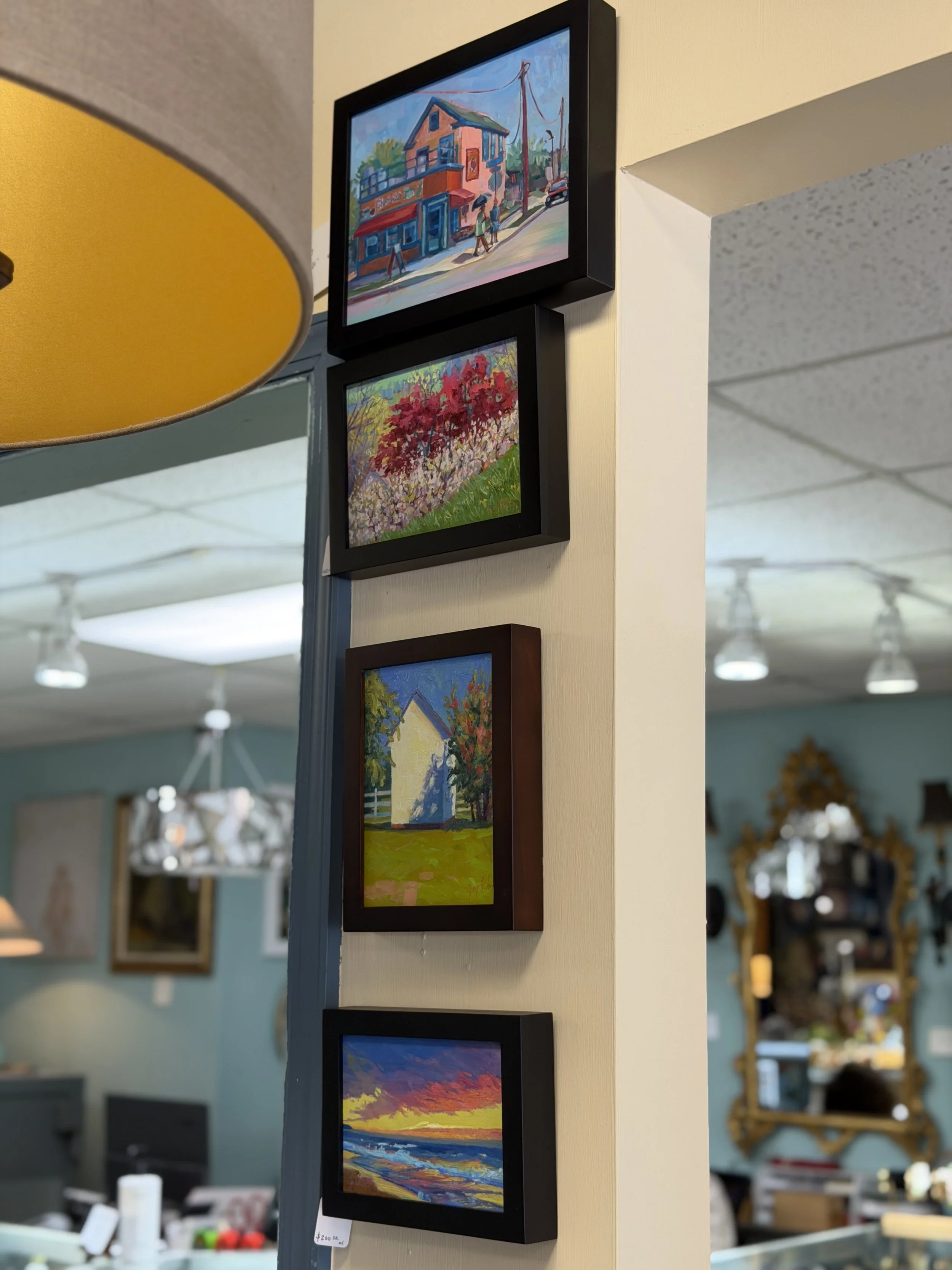 Four framed landscape paintings arranged vertically on a white wall, depicting scenes of a colorful neighborhood, flowering trees, a house, and a sunset over the ocean.