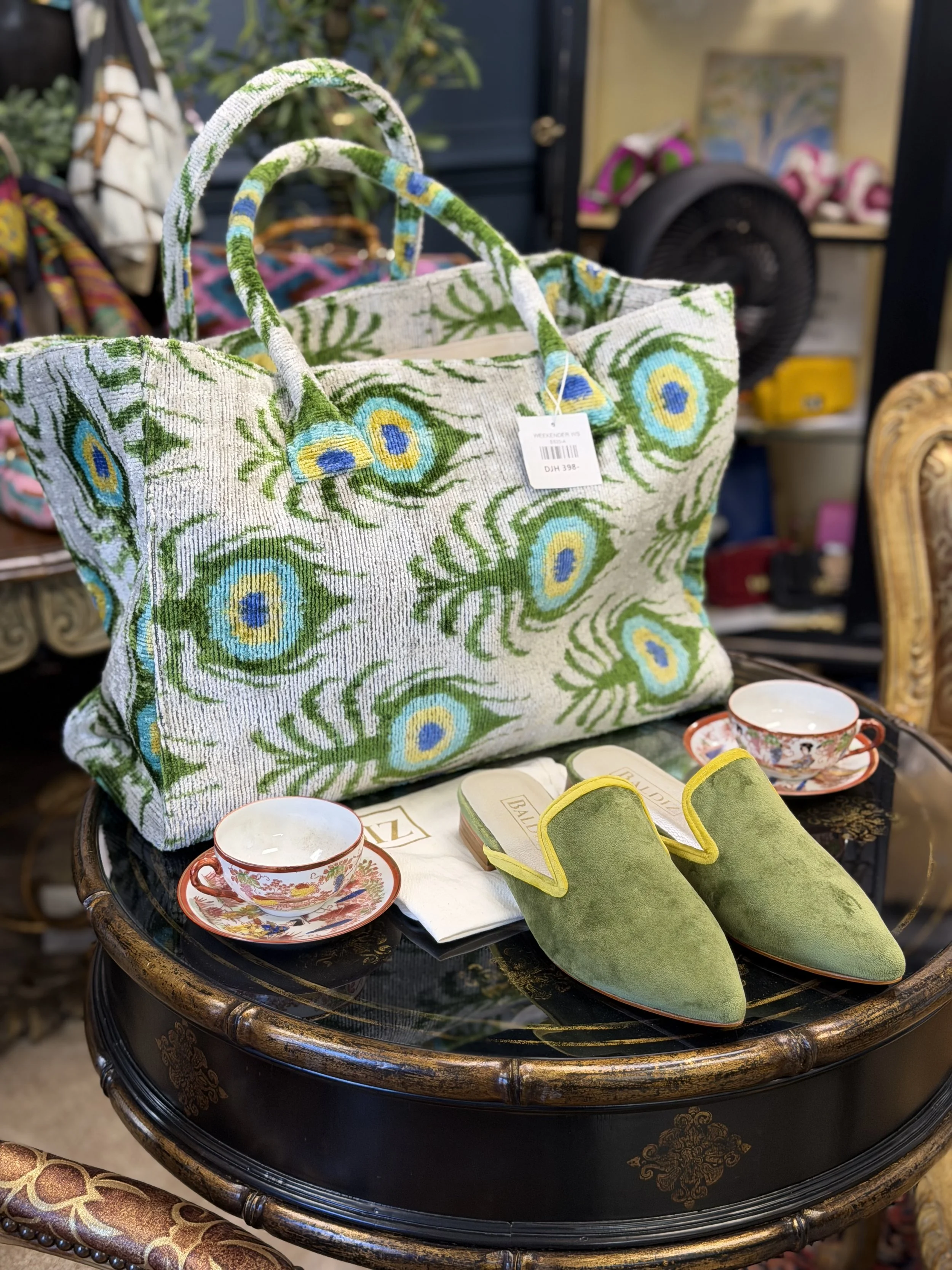 A large woven bag with a floral pattern, green slippers, and two teacups with saucers on a small round table in a vintage shop.