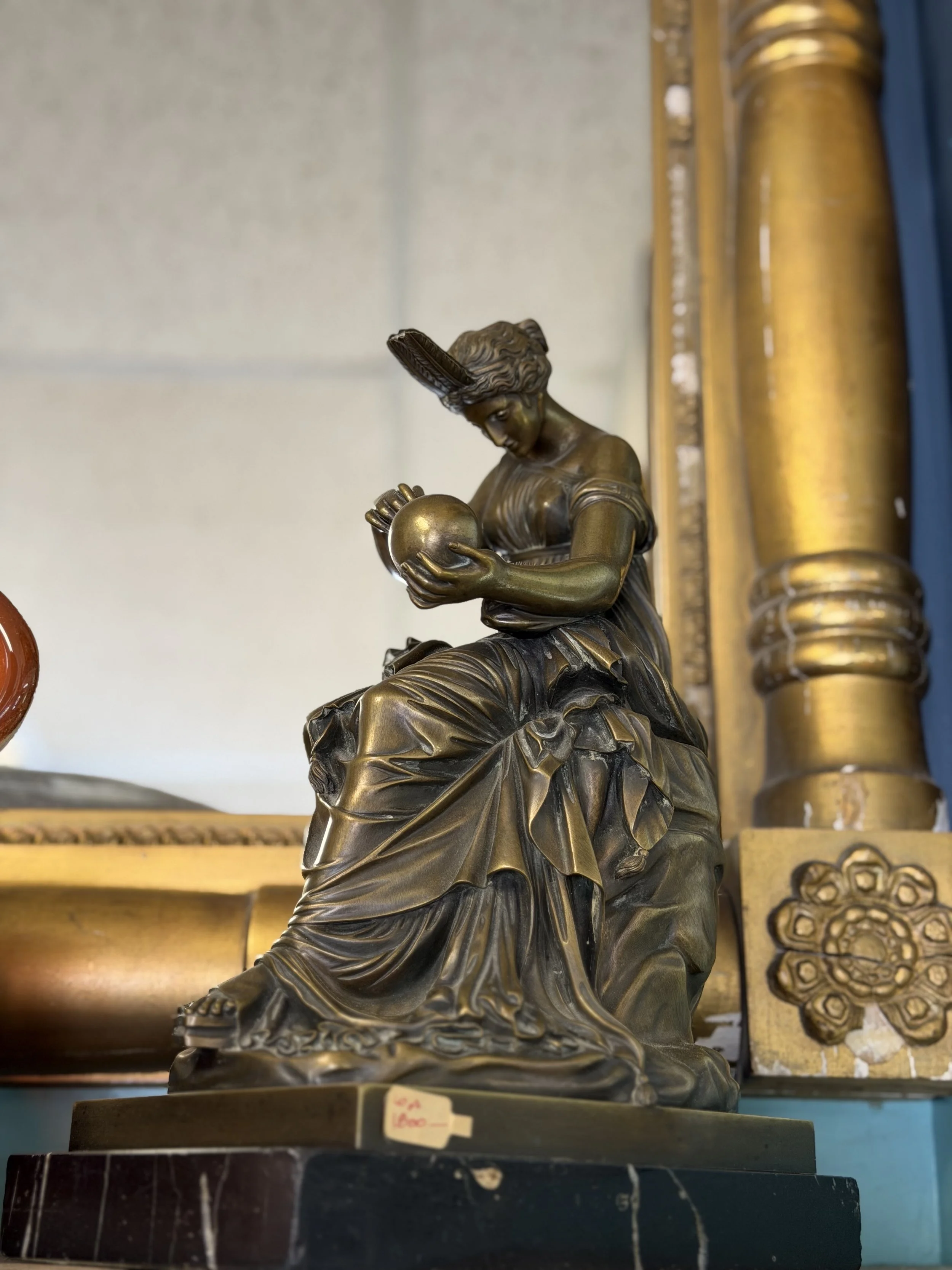 Bronze statue of a woman in classical attire holding a spherical object with a feather in her hair, seated on a decorative base, behind a gold-colored column.