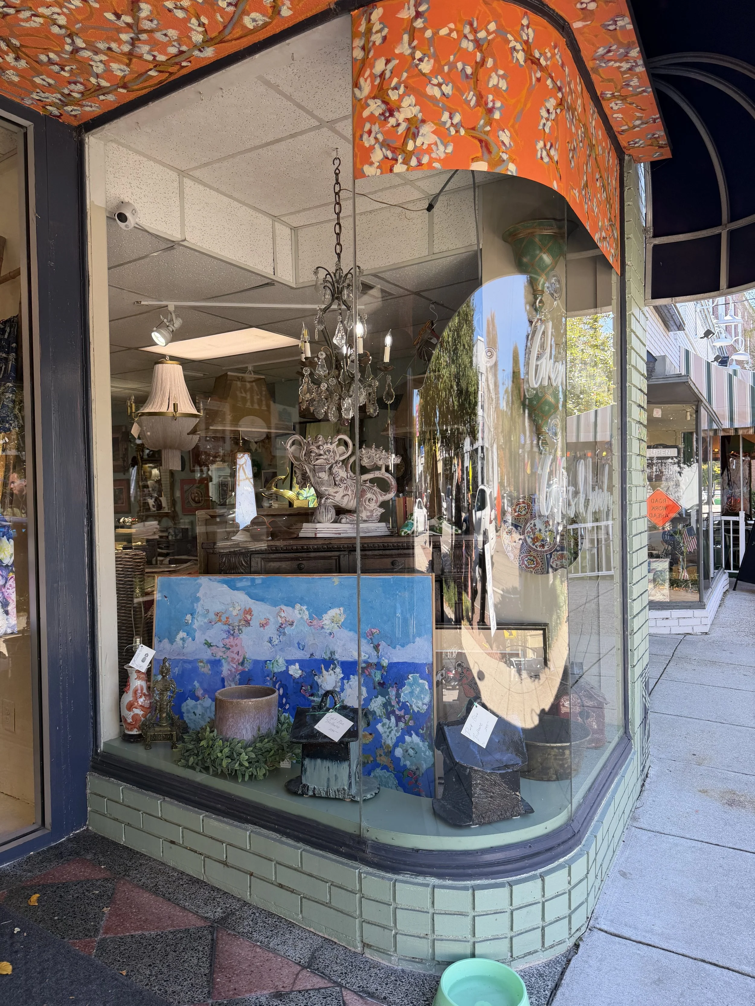 View of an antique shop window displaying various decorative items including a chandelier, lamps, artwork, vases, and other collectibles. The shop has a green brick exterior and reflections of the street and trees are visible on the glass.