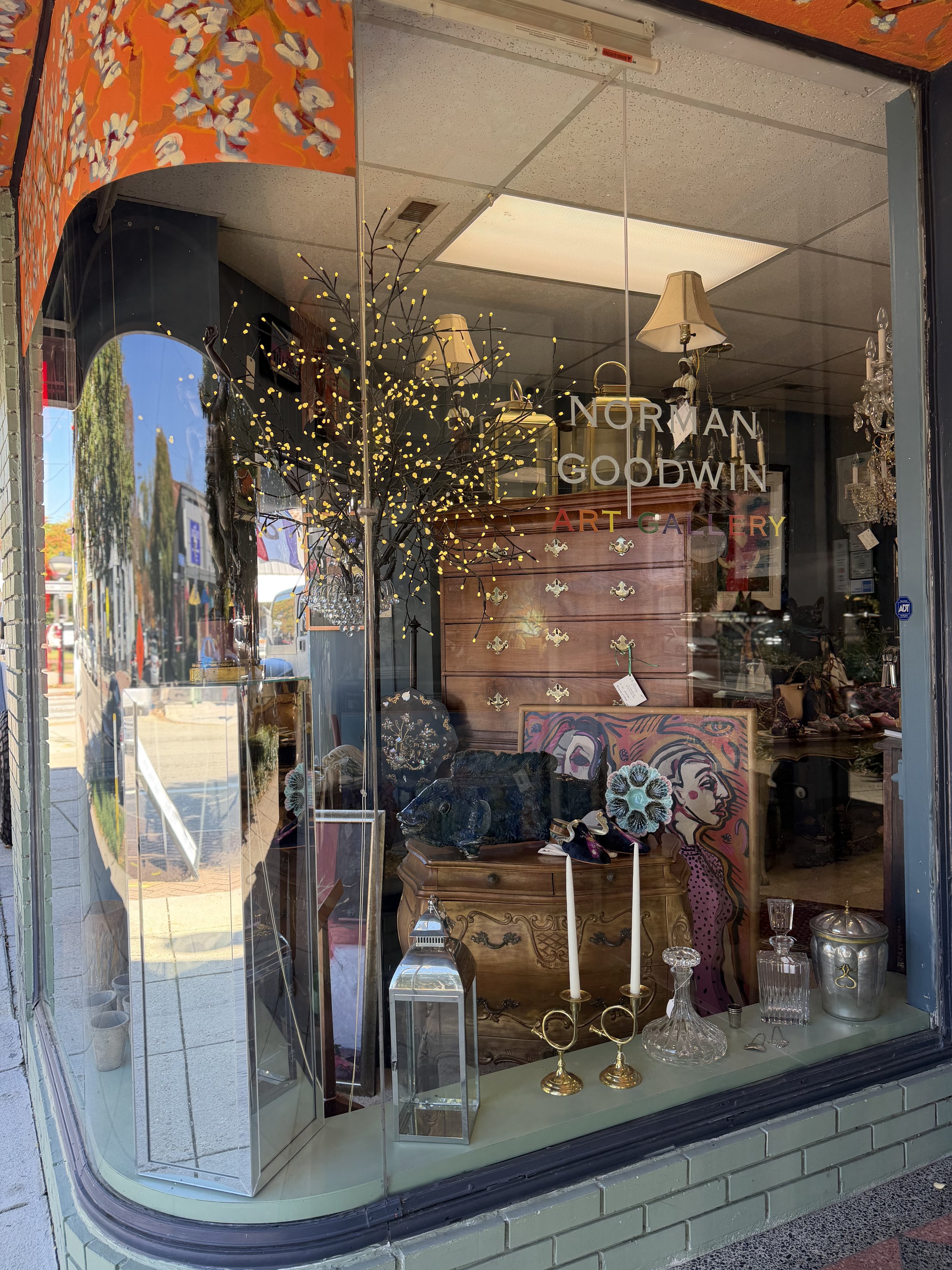 A storefront window display featuring various decorative items including a lit-up artificial tree, a black sculpture of a bear, vintage-style liquor bottles, brass candlesticks with candles, a framed artwork of a woman's face, and a wooden chest of d