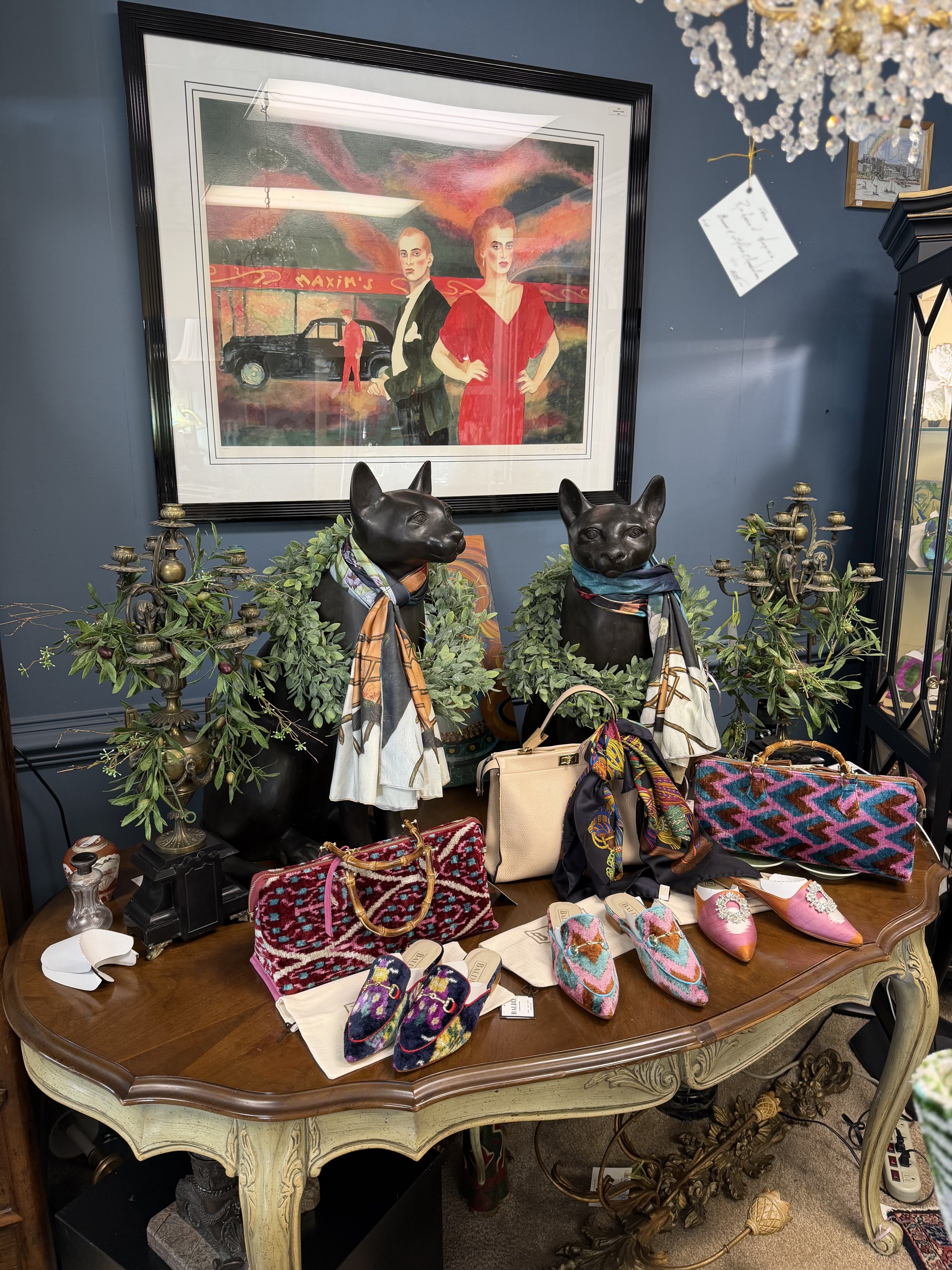 A decorative display with two black dog sculptures wearing scarves, surrounded by handbags and slippers on a vintage-style wooden table, with an art print of two women and a car hanging on a dark blue wall behind them.