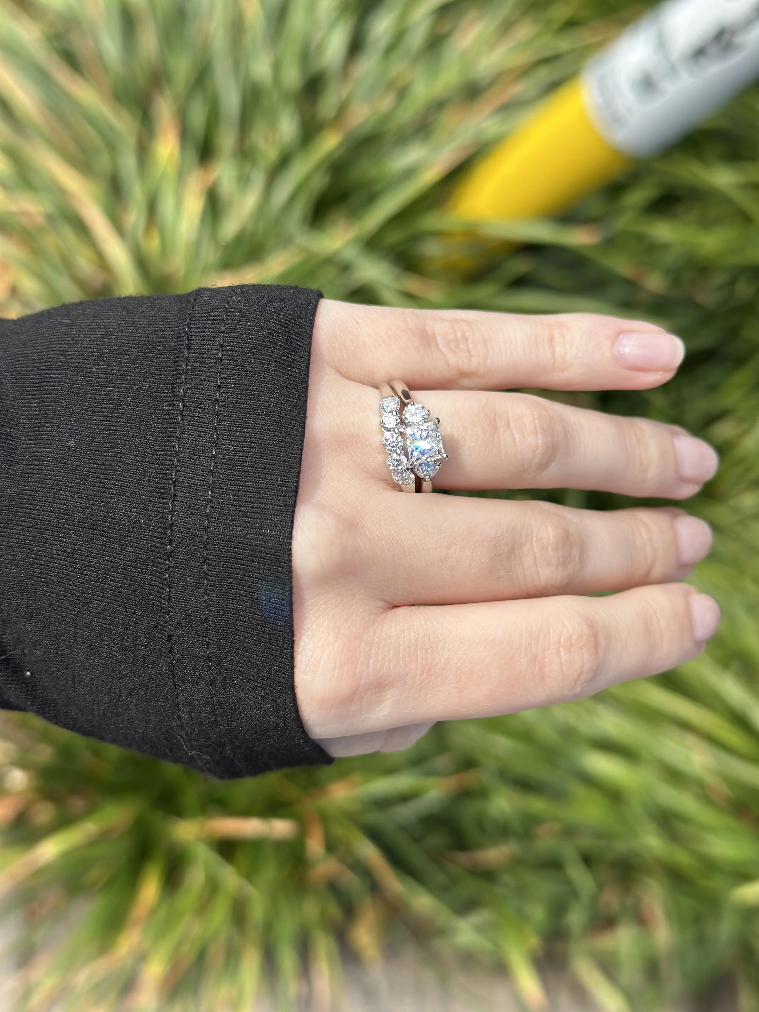 A person's hand wearing a silver ring with multiple large and small clear gemstones, held against a blurry green background of foliage.